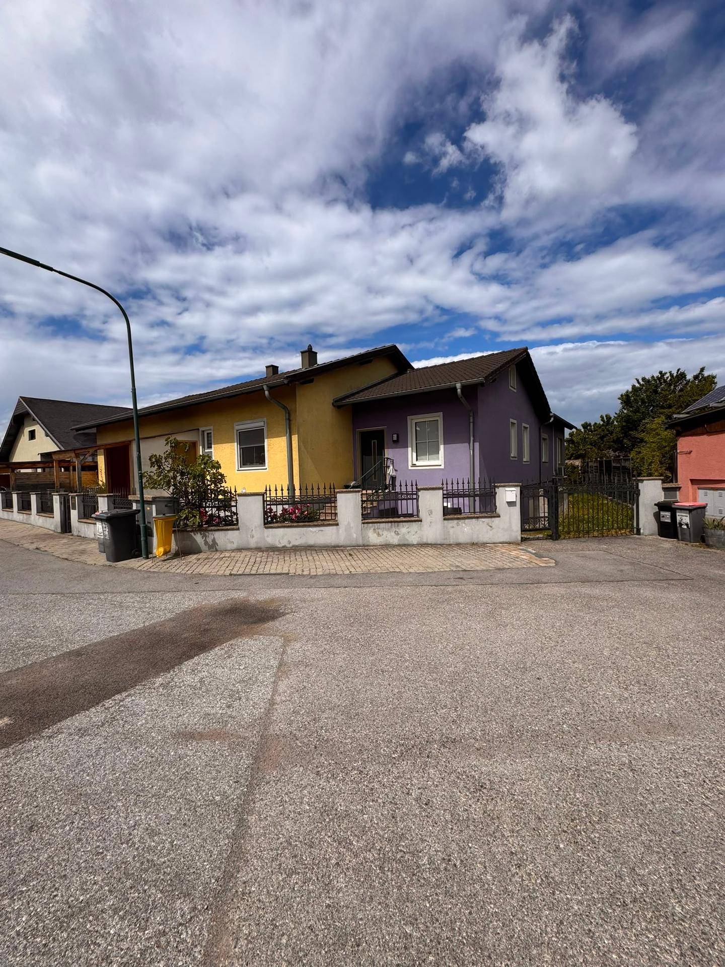 Einfamilienhaus mit gelber und lila Fassade, umgeben von einem Zaun und Bäumen.