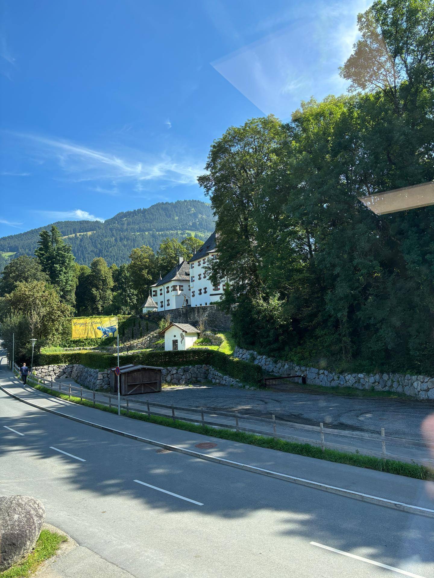 Grüne Umgebung mit Bergen im Hintergrund, einer Straße und einem historischen Gebäude unter blauem Himmel.