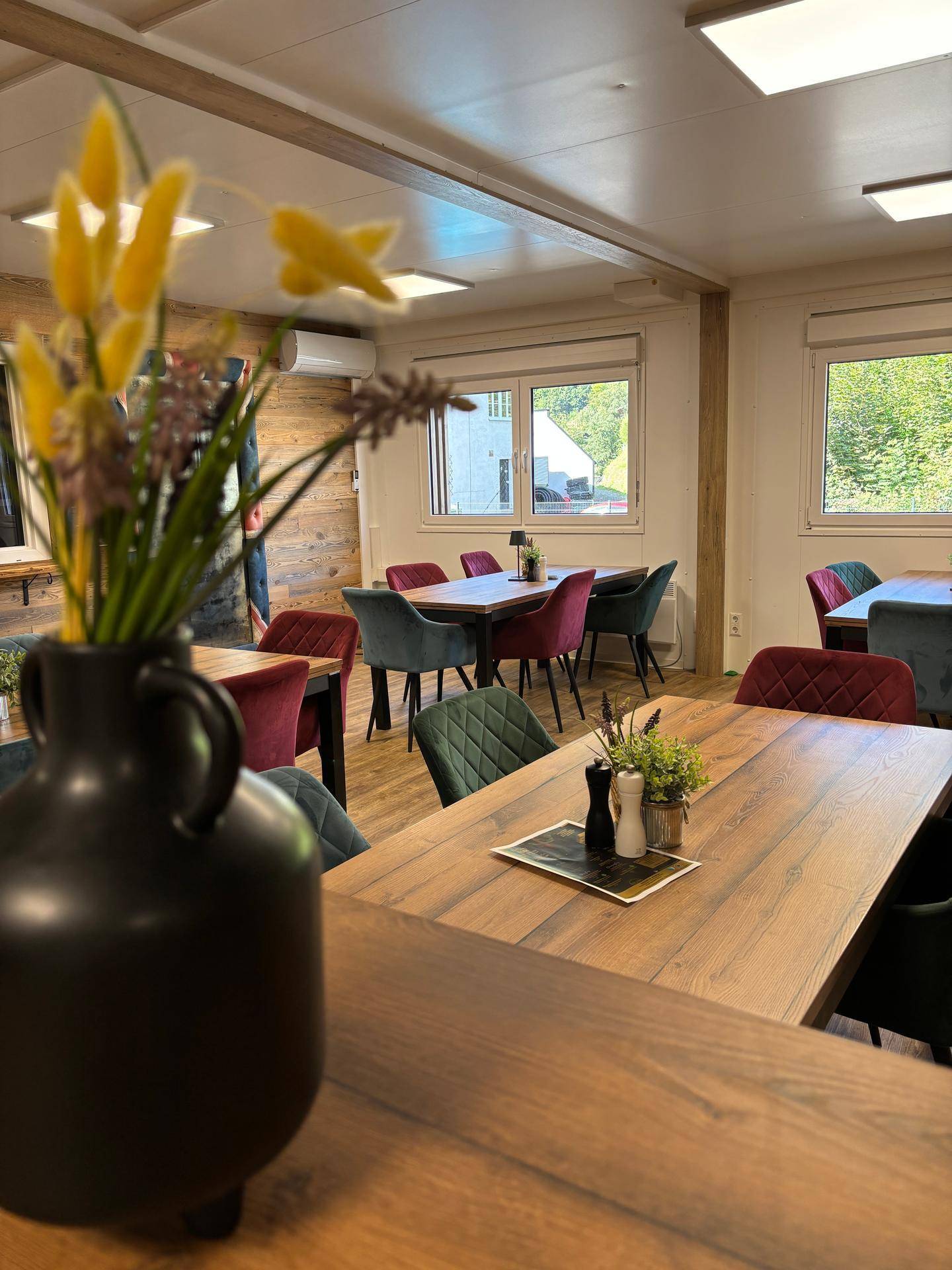 Heller Essbereich mit Holztischen, farbigen Stühlen und Fenstern mit Blick auf die grüne Umgebung.