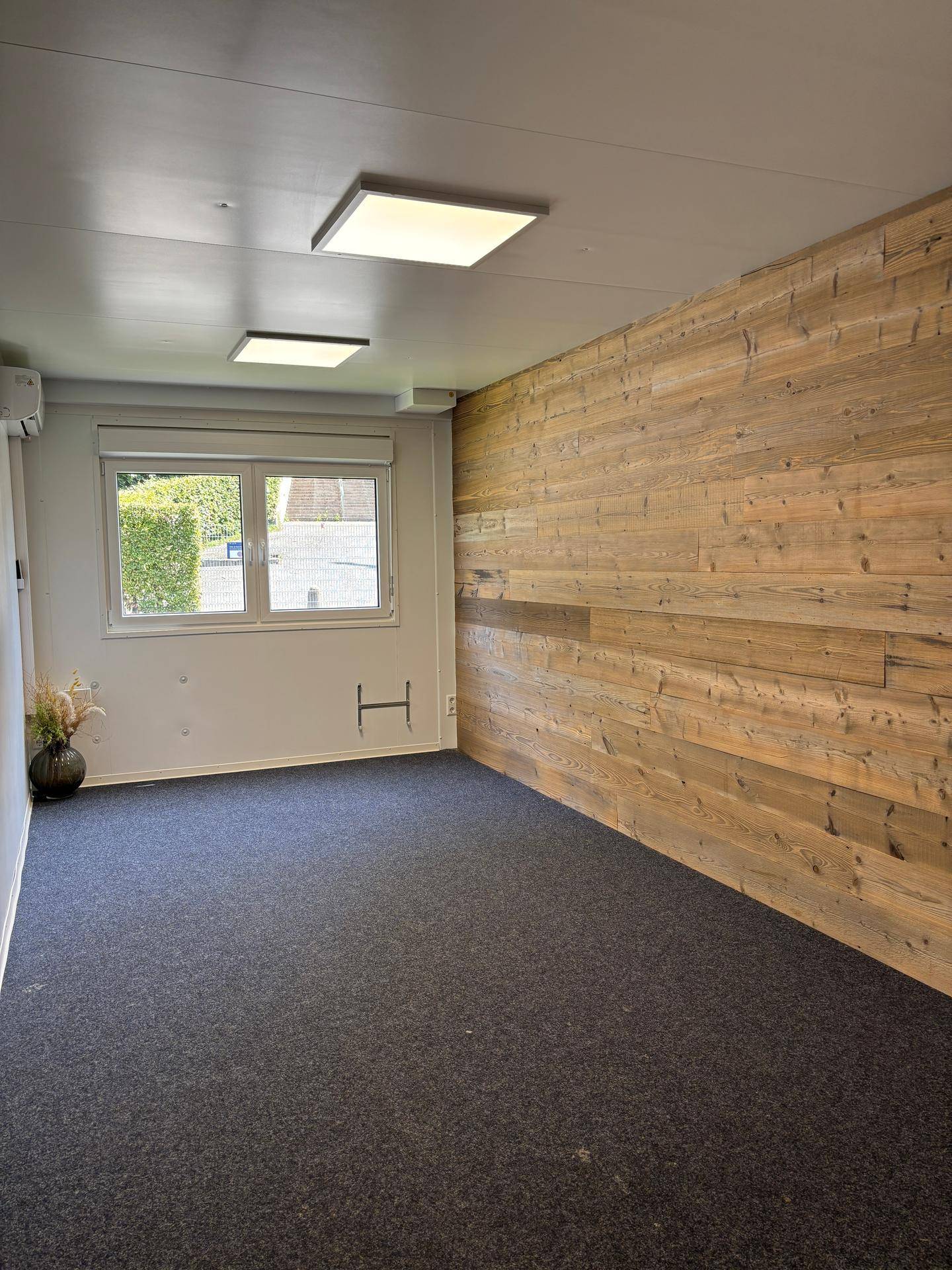 Leeres Büro mit Holzwand, dunklem Teppichboden und Fenster, das natürliches Licht hereinlässt.