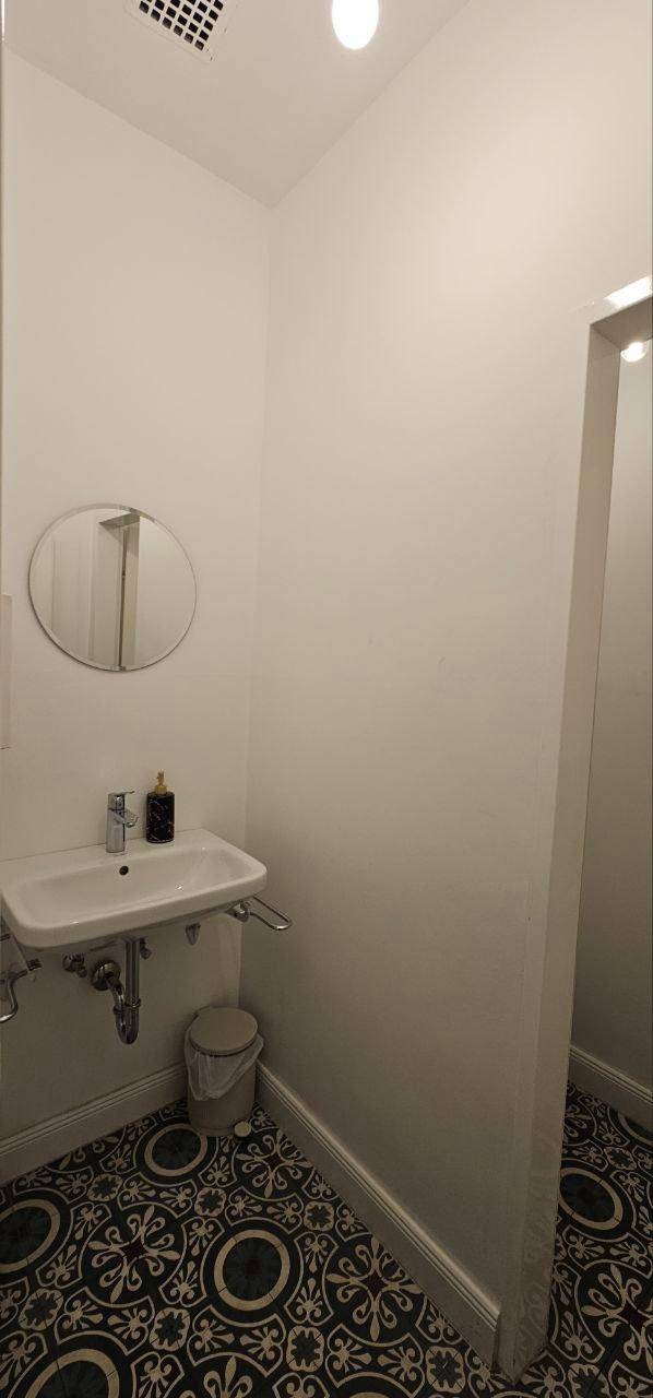 Waschbeckenbereich in einer kleinen Toilette mit rundem Spiegel und gemusterten Bodenfliesen.