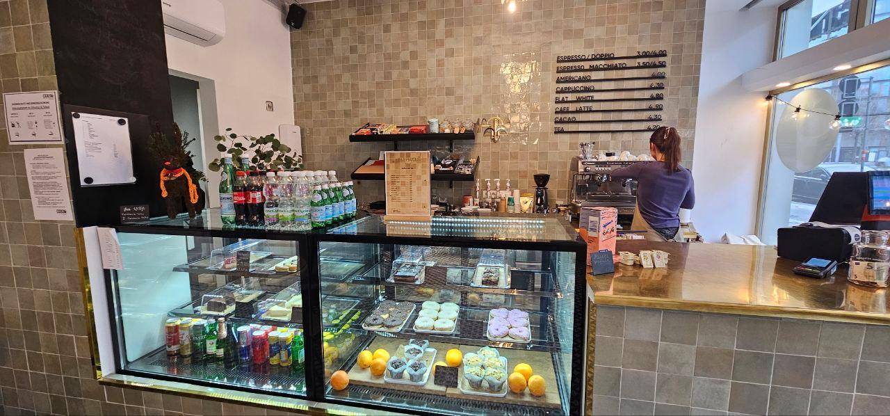 Thekenbereich eines Cafés mit Kuchenvitrine, Getränken und einer Barista bei der Arbeit.