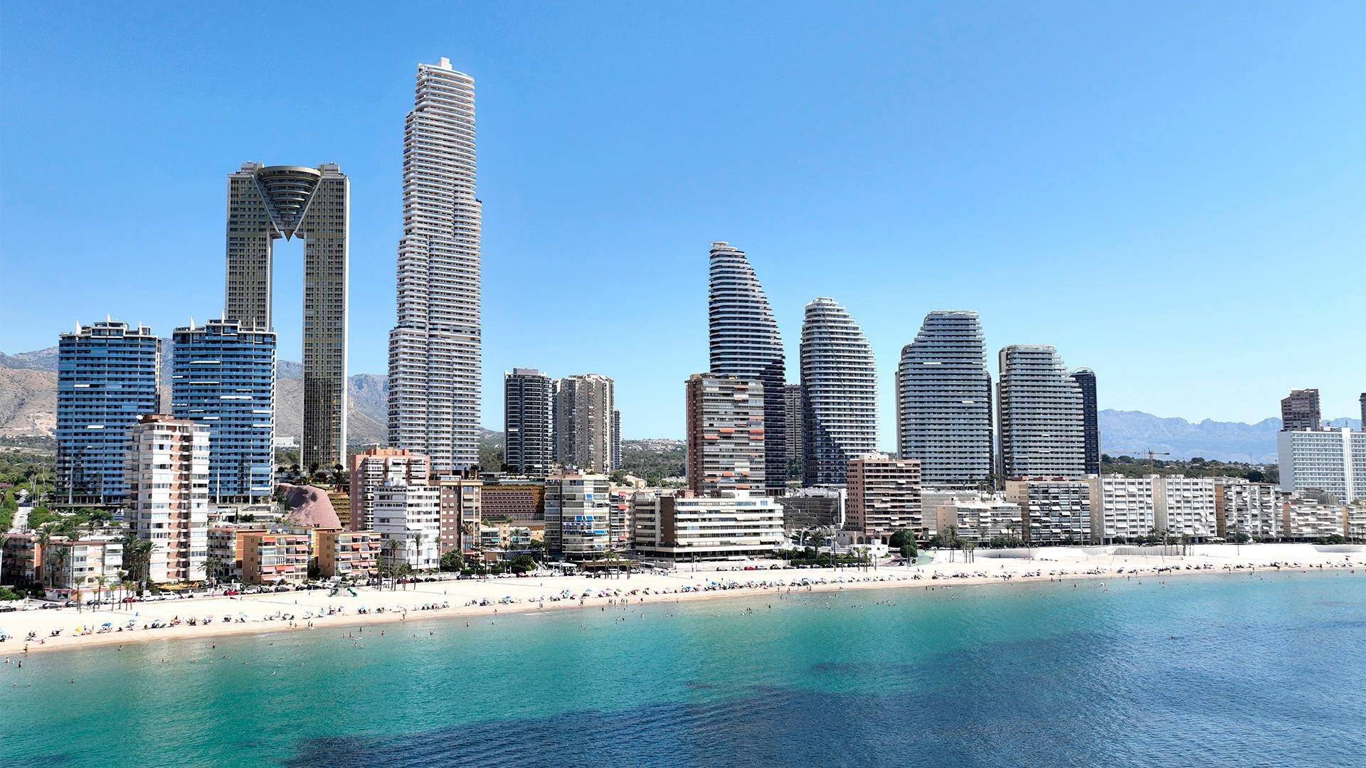 Weitläufiger Strand mit Stadtpanorama, modernen Hochhäusern und klarem blauen Meer unter strahlendem Himmel.
