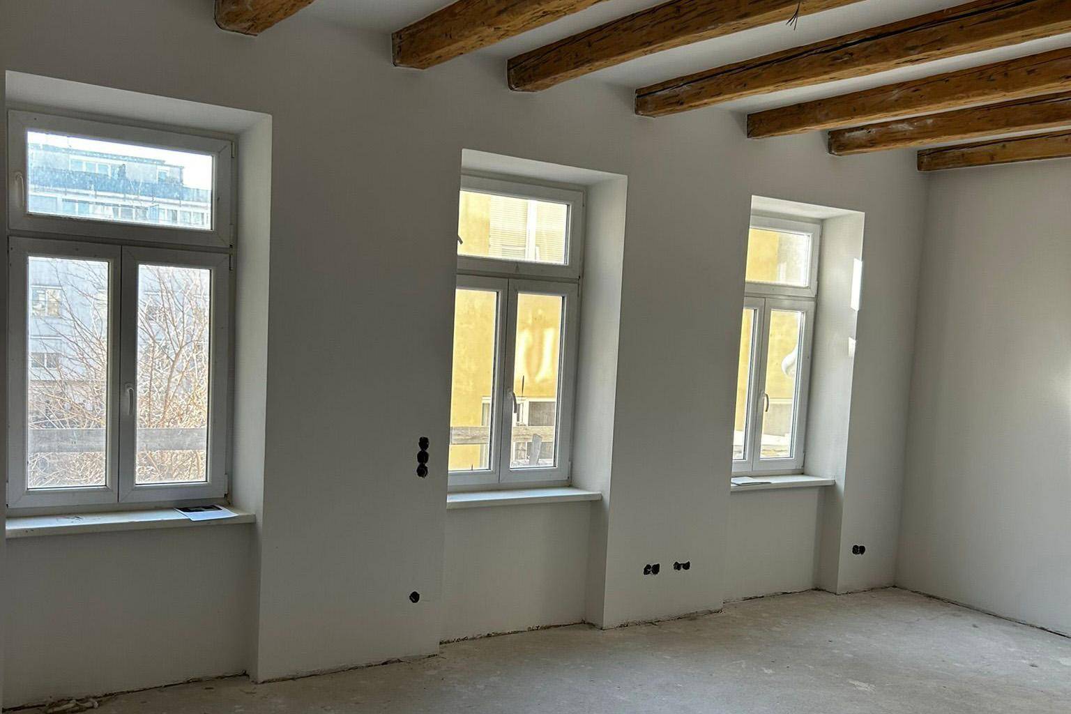 Heller Rohbau mit drei Fenstern und sichtbaren Holzbalken, wartet auf den Innenausbau.