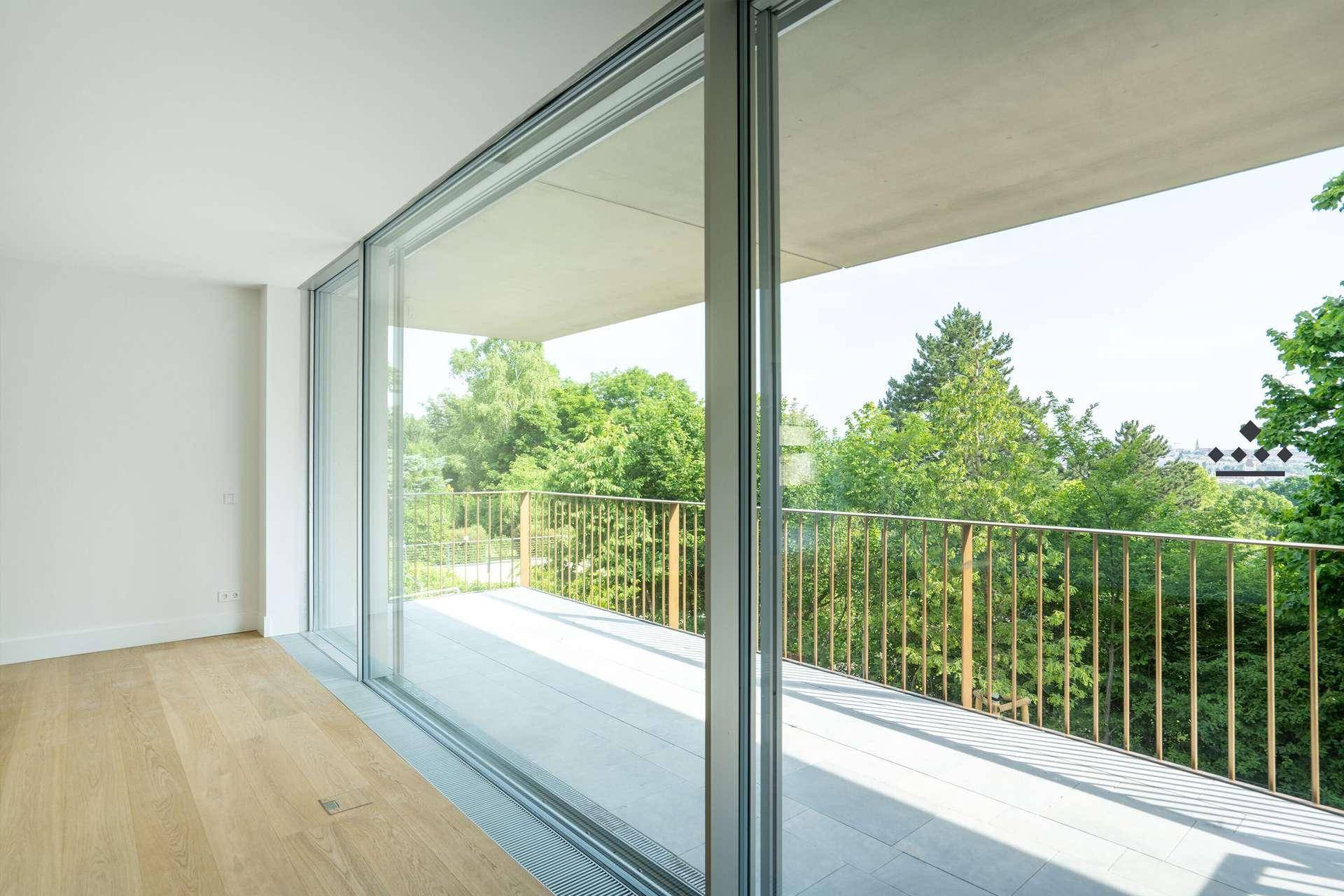Heller Raum mit Holzboden und großen Schiebetüren, die zu einem Balkon mit Baumblick führen.