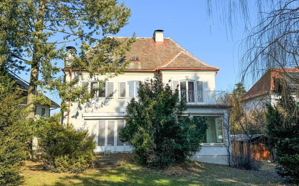 Klassisches Wohnhaus mit rotem Ziegeldach, Balkon und traditionellen Fensterläden umgeben von Grün.