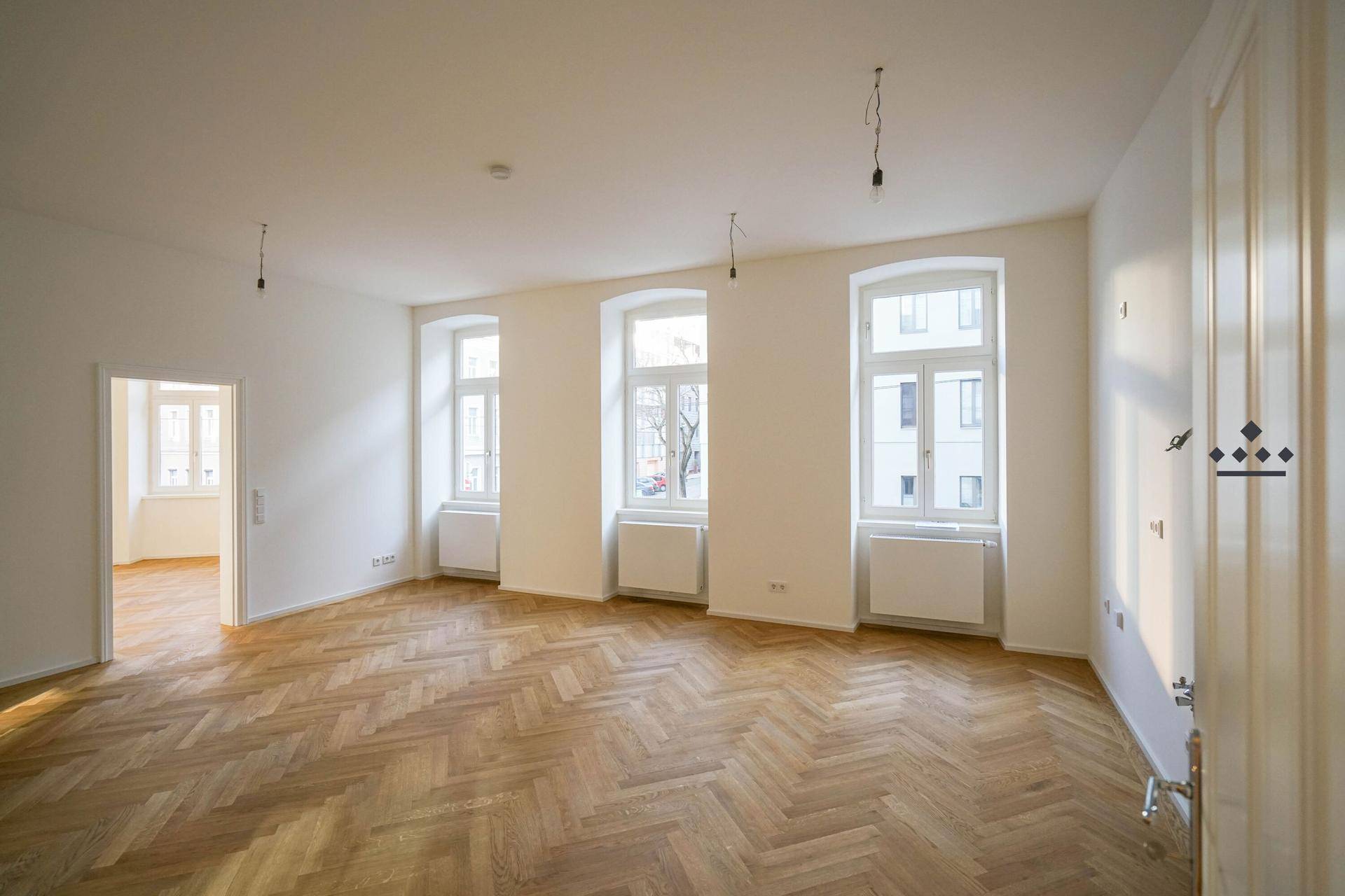 Geräumiger, heller Raum mit Fischgrätparkett, hohen Fenstern und weiß gestrichenen Wänden.