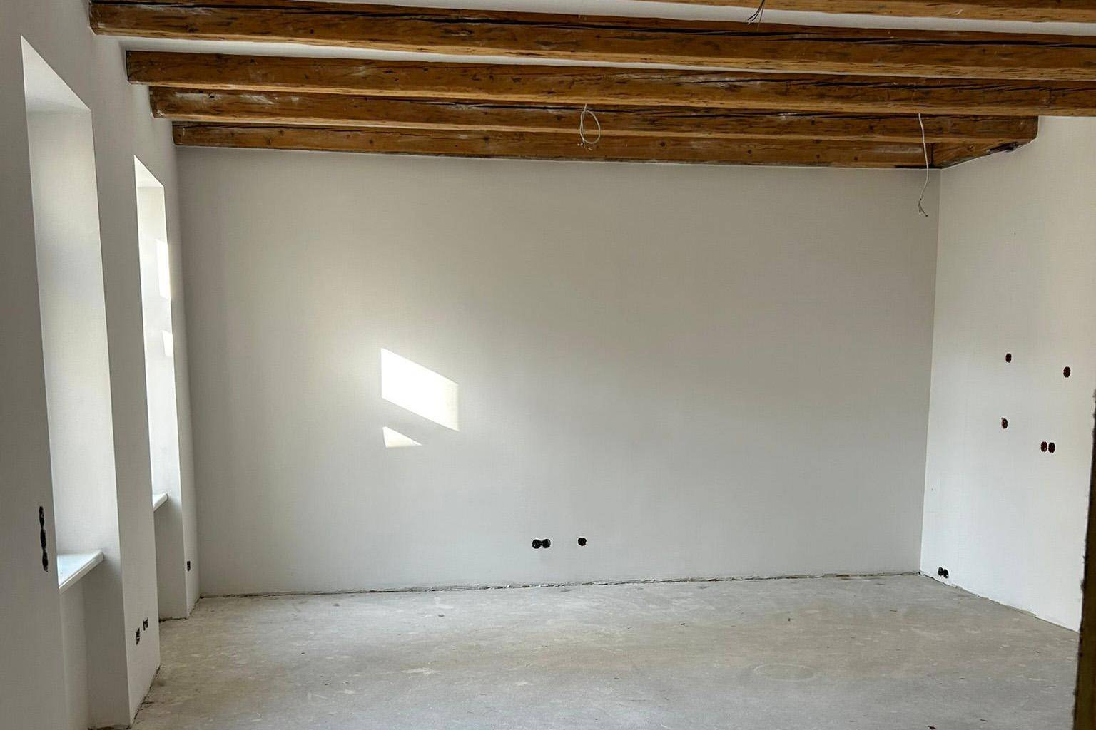 Unfertiger Raum mit sichtbaren Holzbalken und neu verputzten Wänden, bereit für den Ausbau.