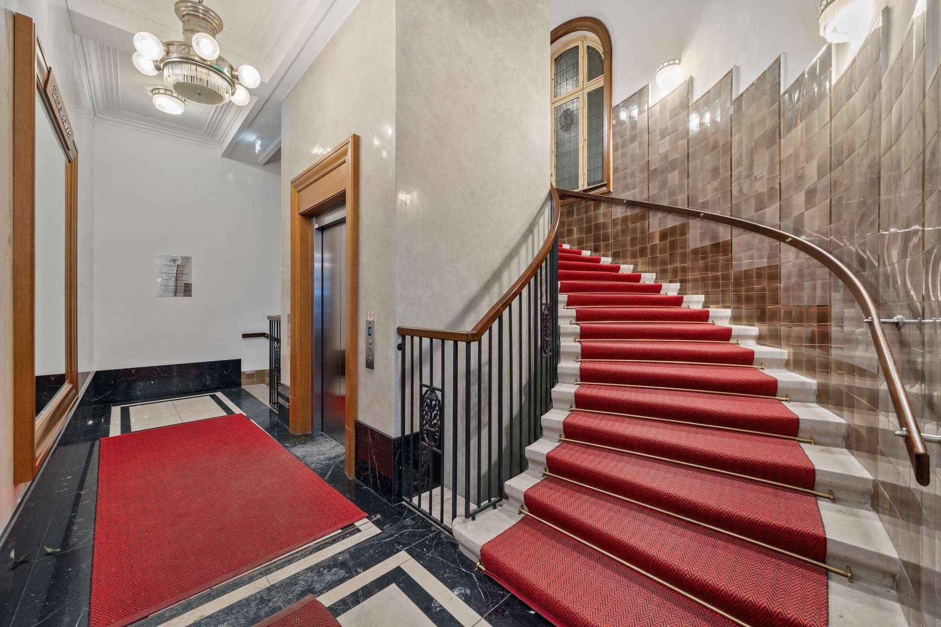 Elegantes Treppenhaus mit rotem Teppich, Marmorverkleidung und einem Aufzug im Hintergrund.