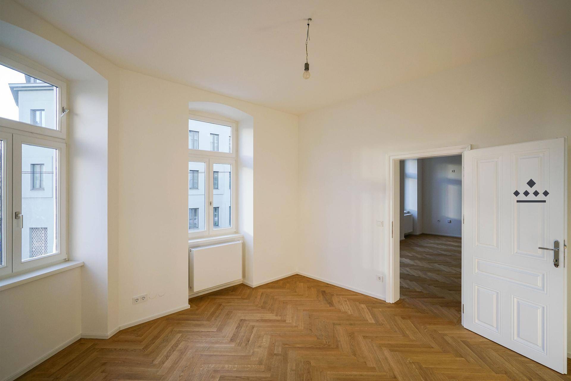 Lichtdurchfluteter Raum mit Fischgrätparkett und Zugang zu einem weiteren Zimmer.