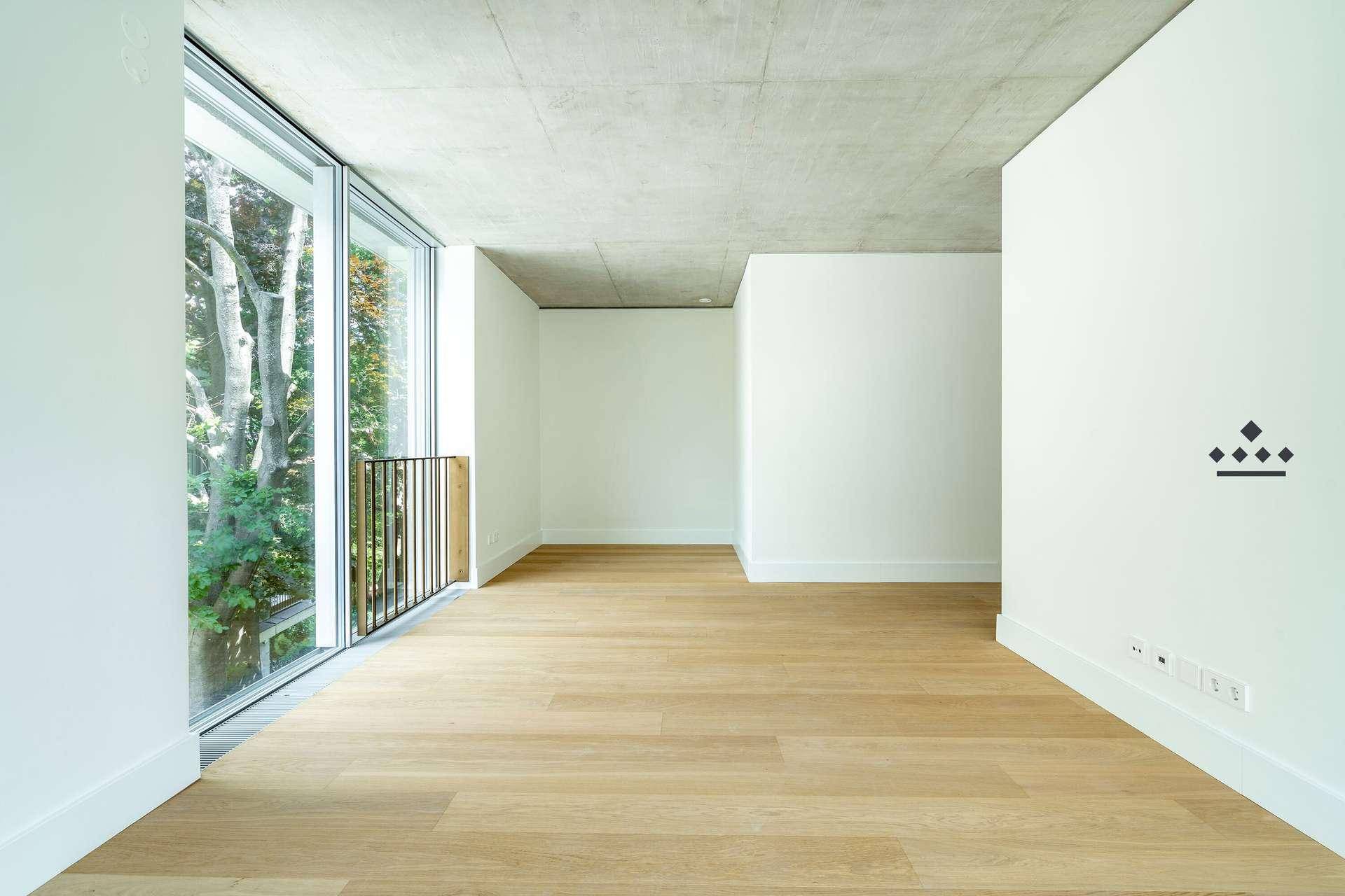 Heller, leerer Raum mit hohen Decken, Holzboden und bodentiefen Fenstern mit Blick ins Grüne.