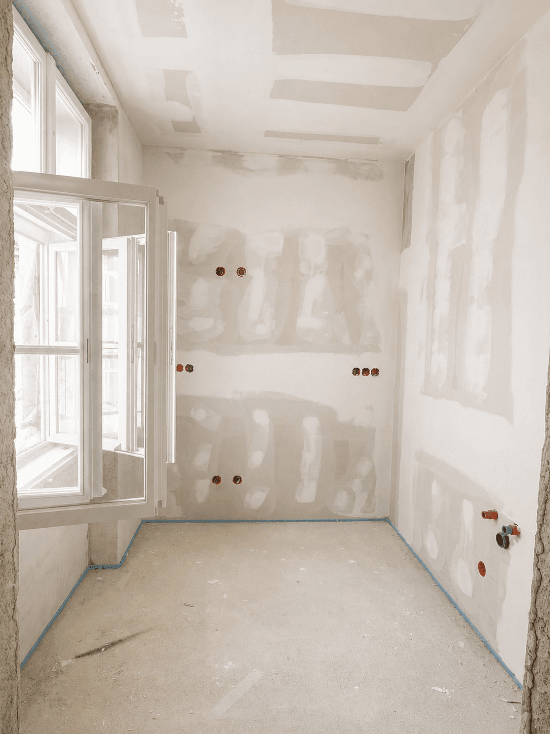 Unfertiger Raum mit Fenster und vorbereiteten Anschlüssen für Sanitärinstallationen.