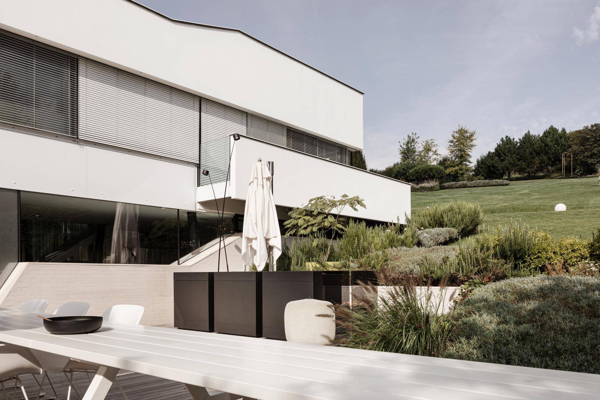 Große Terrasse mit Esstisch und Sonnenschirm, umgeben von einer gepflegten Gartenlandschaft und Hügeln.