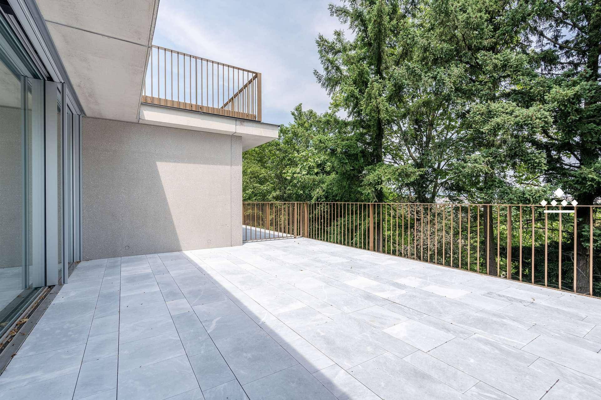 Große, geflieste Terrasse mit Metallgeländer und Blick auf grüne Bäume.