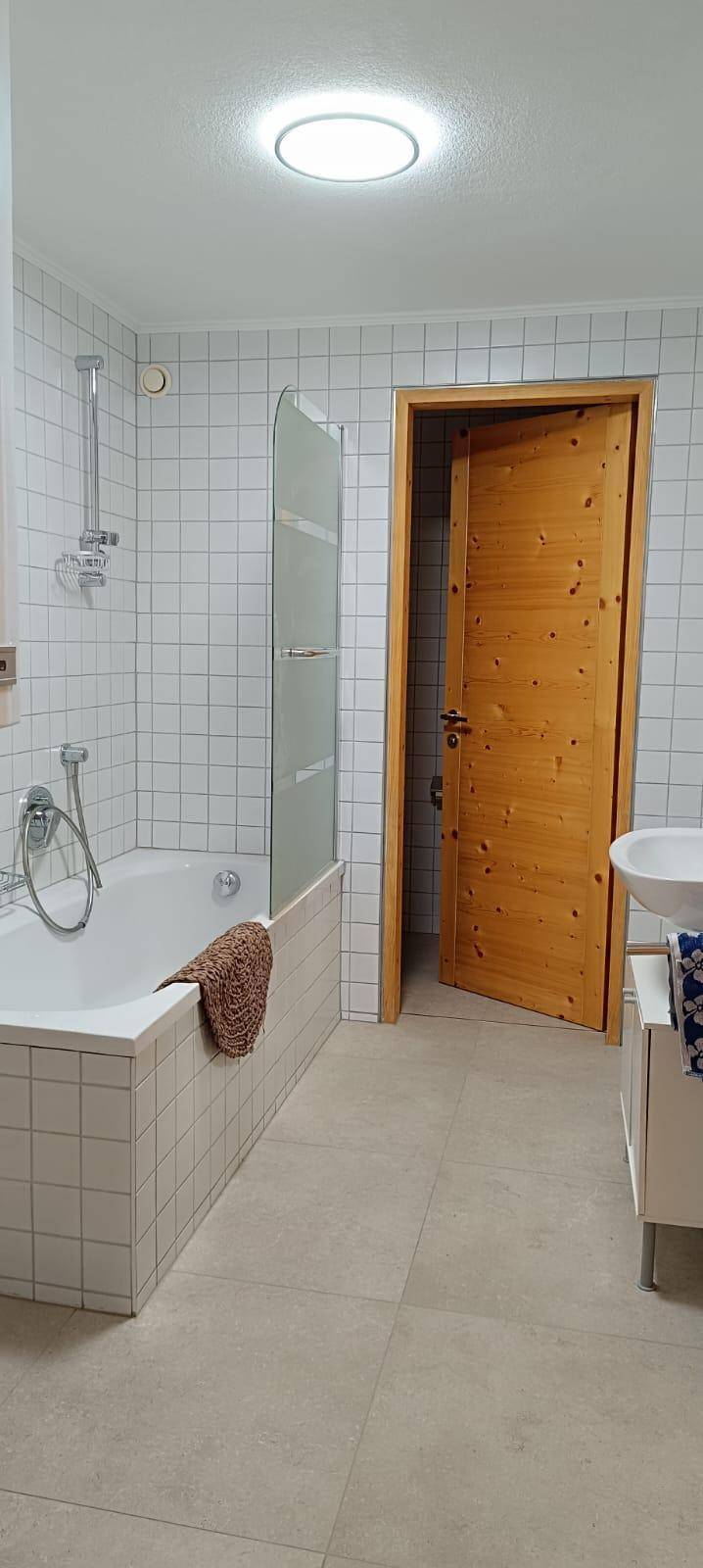 Badezimmer mit Badewanne, Duschvorrichtung und weißen Fliesen, ausgestattet mit einer Holztür.