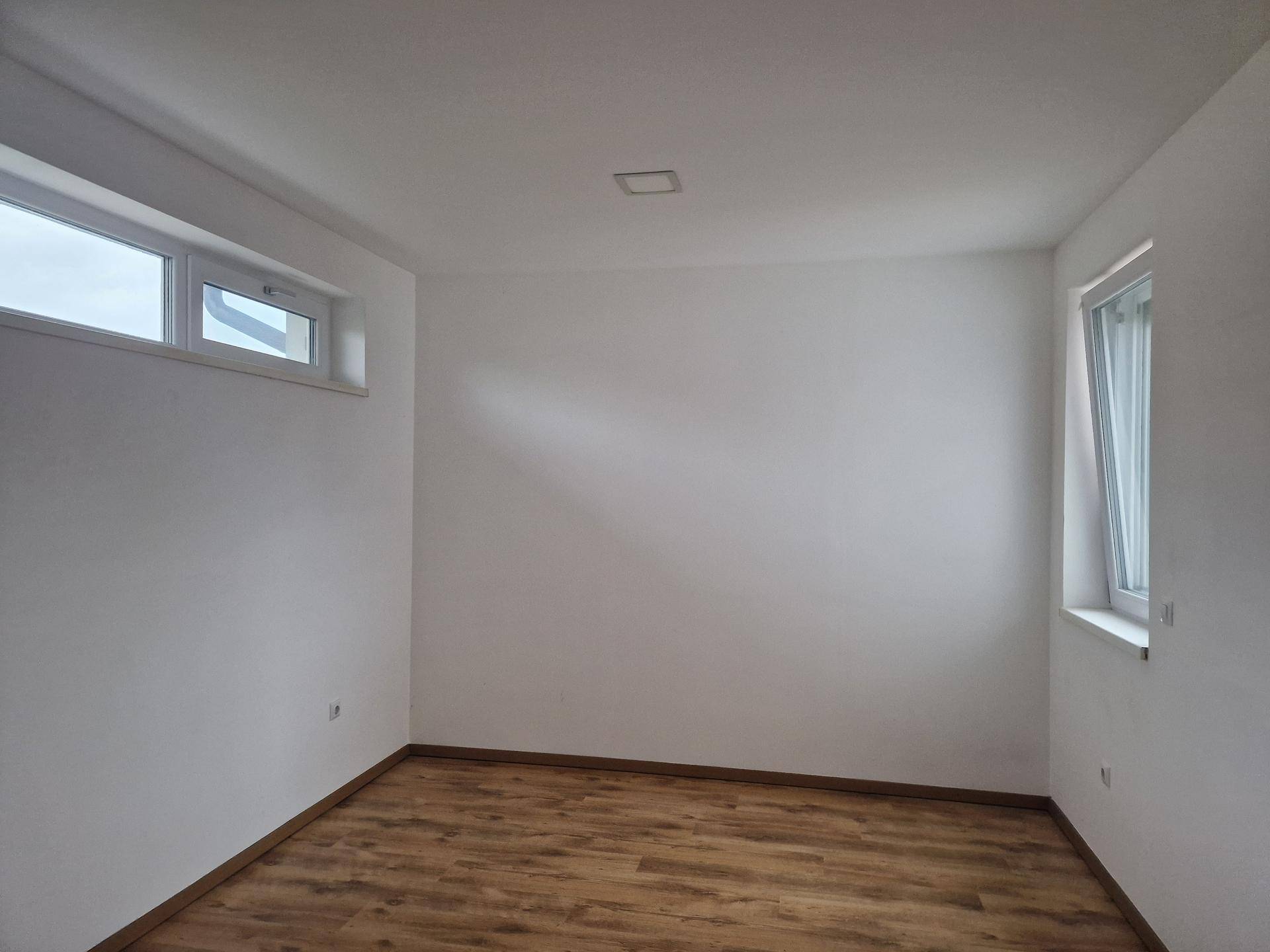 Leeres Zimmer mit zwei Fenstern, weiß gestrichenen Wänden und modernem Laminatboden.
