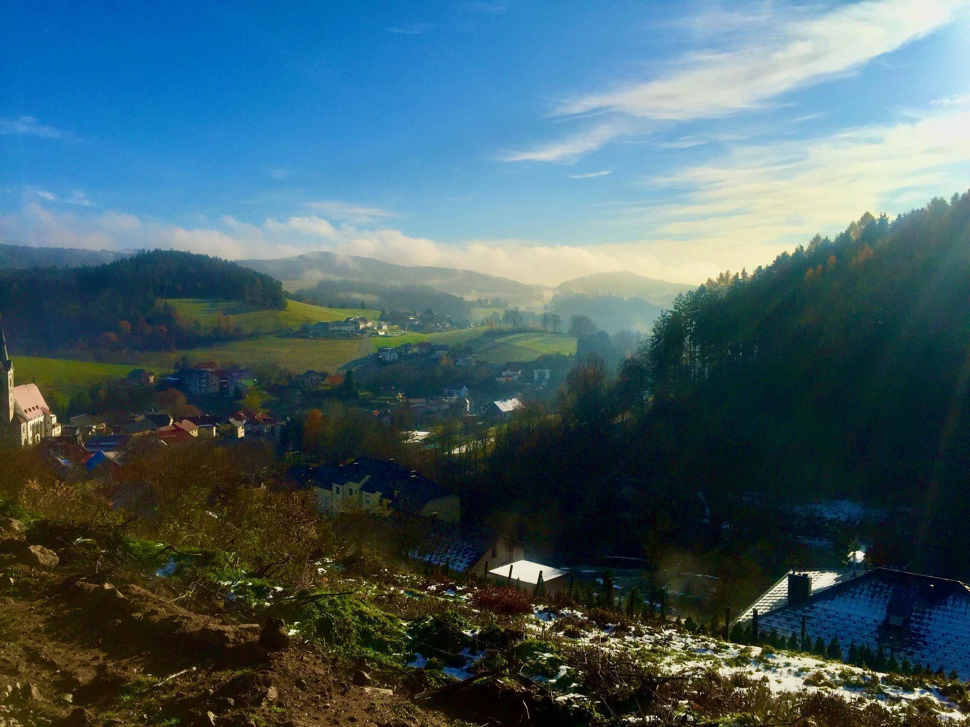 Panoramablick auf ein idyllisches Dorf in einer hügeligen Landschaft mit ersten Schneespuren und Sonnenstrahlen.