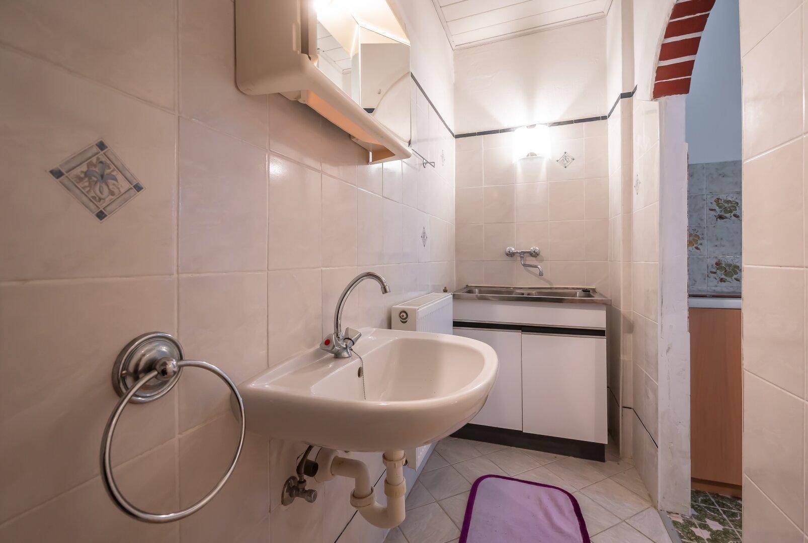 Badezimmer mit Waschbecken, Spiegelschrank und Fliesen, angrenzend an die Küche.