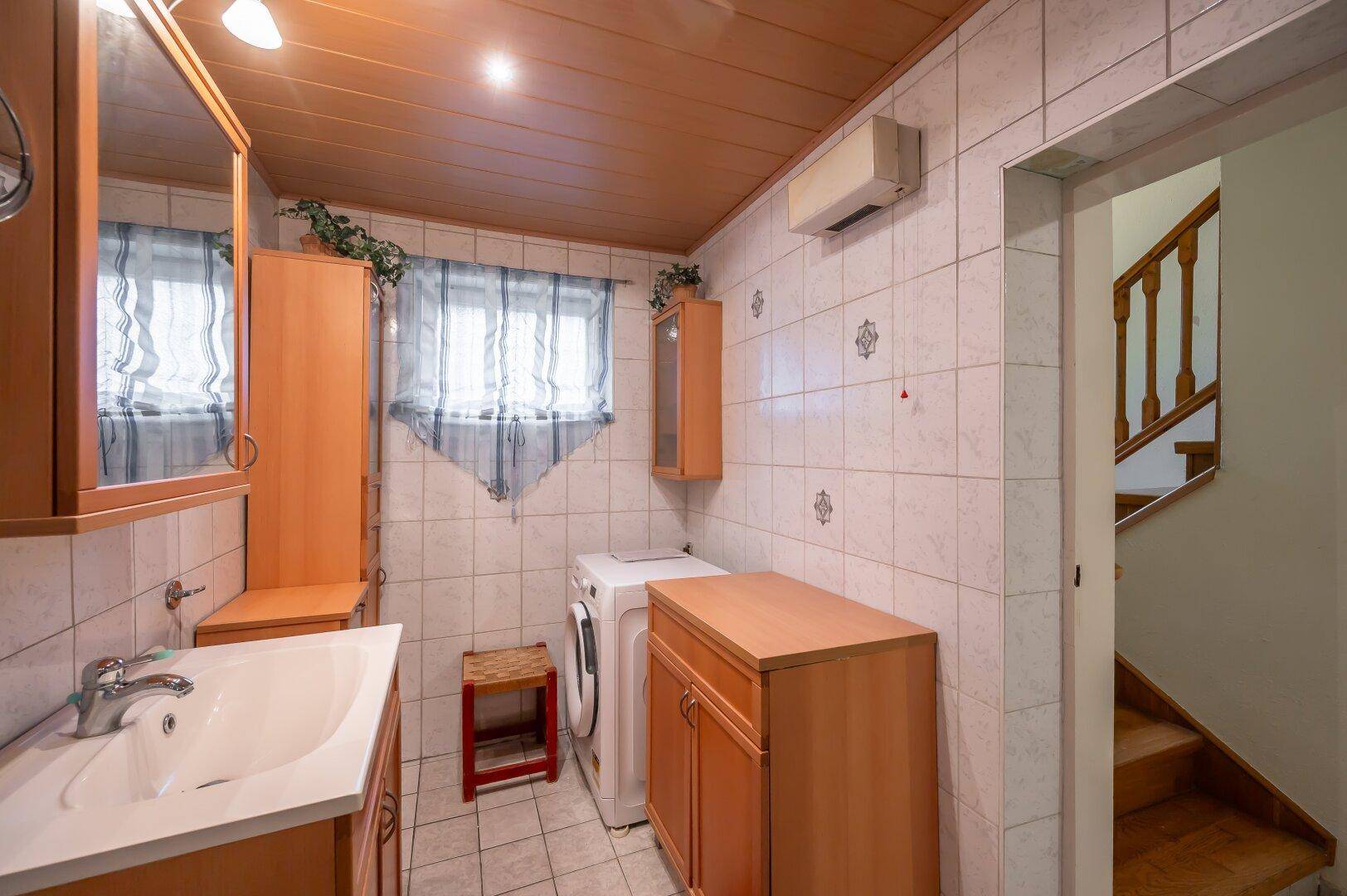 Badezimmer mit Waschbecken, Waschmaschine und Fliesenwänden, unter einer Holzdecke.