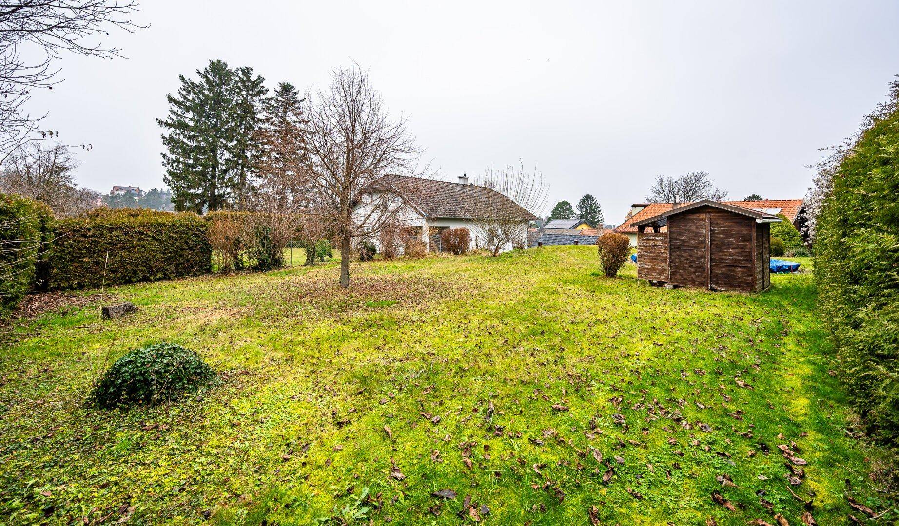 Weitläufiger Garten mit Rasenfläche und einem kleinen Gartenhaus, umgeben von Bäumen.