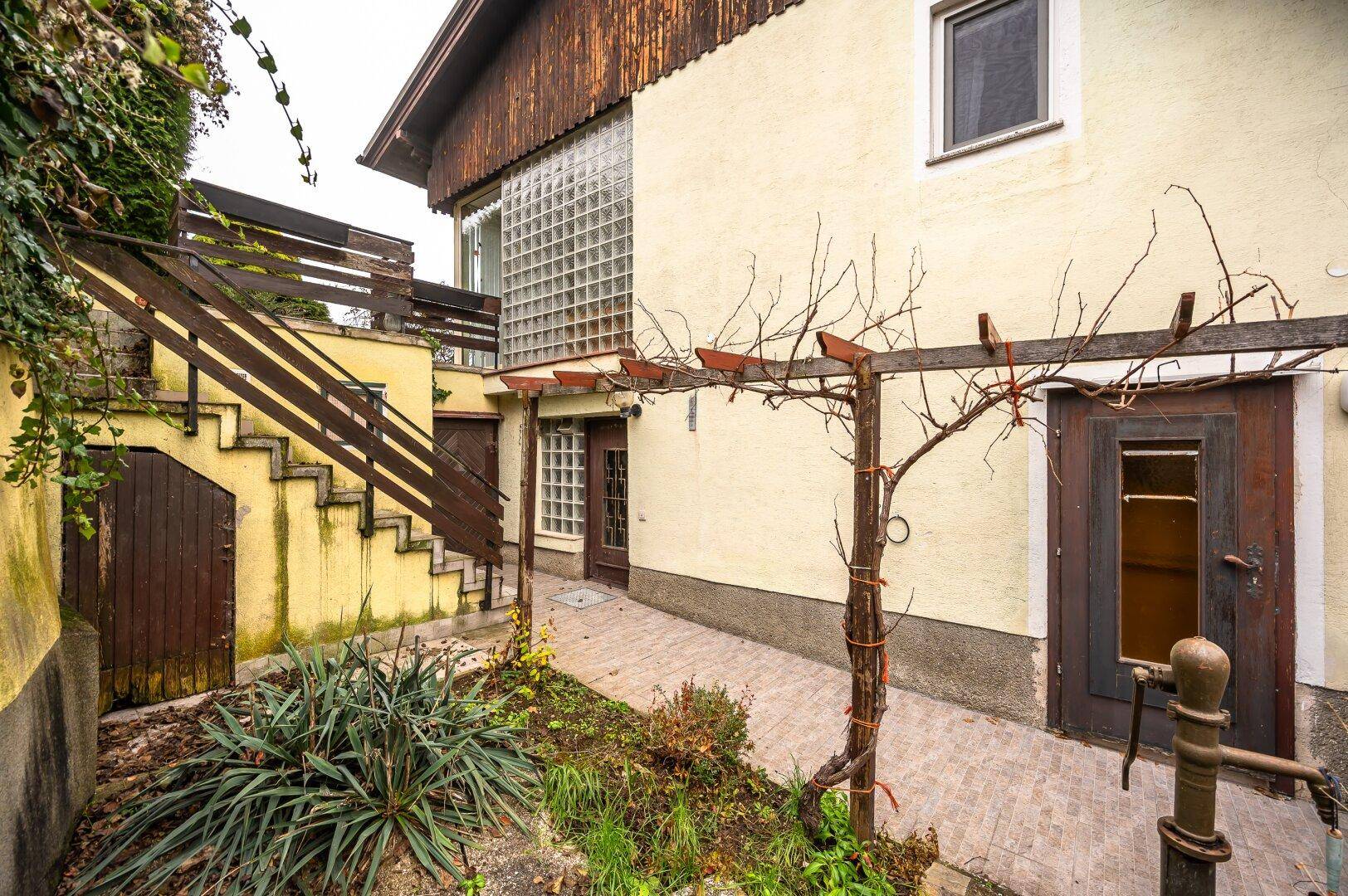 Seitlicher Eingangsbereich mit Holztreppe, gepflastertem Hof und einer Pergola-Struktur im Garten.