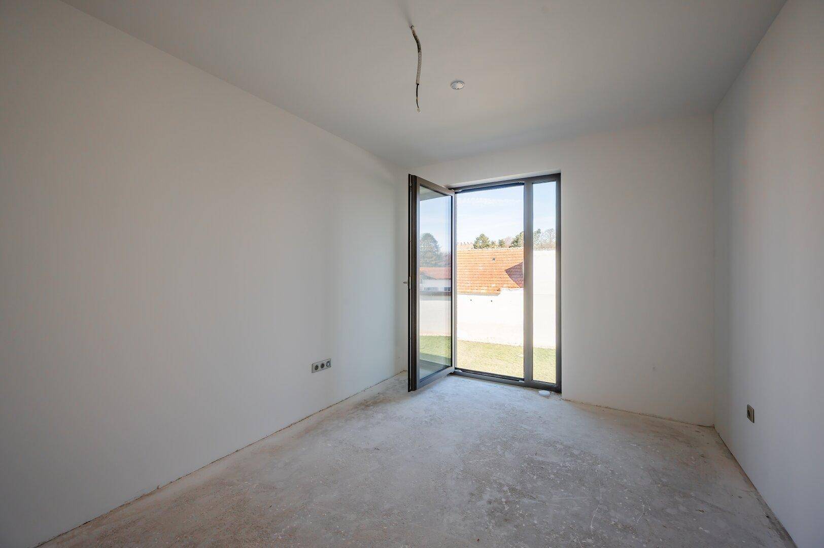 Helles Schlafzimmer im Rohbau mit Zugang zu einem kleinen Außenbereich und Betonboden.