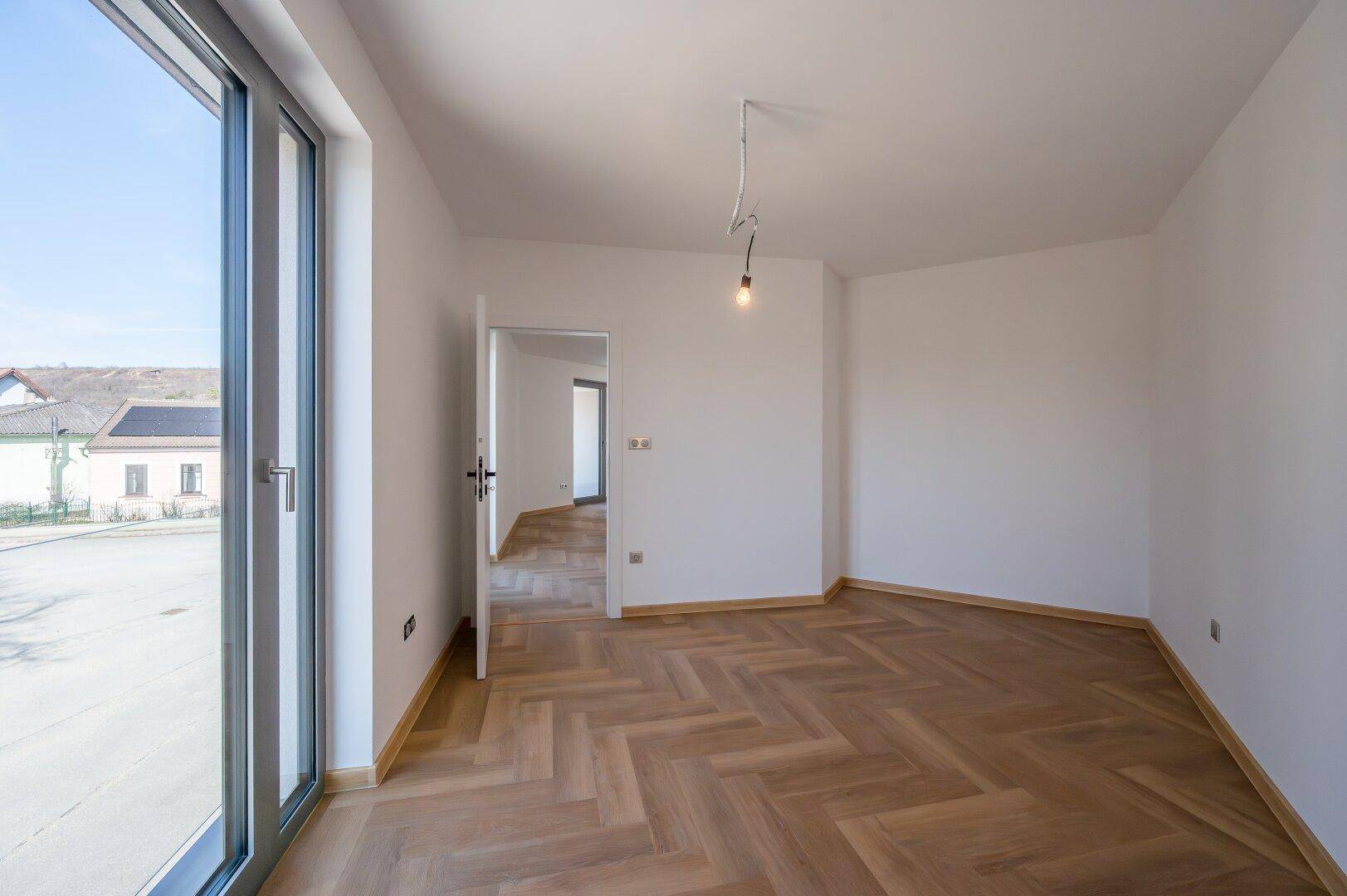 Lichtdurchfluteter Raum mit Fischgrätparkett und Zugang zu einem weiteren Zimmer sowie zur Terrasse.