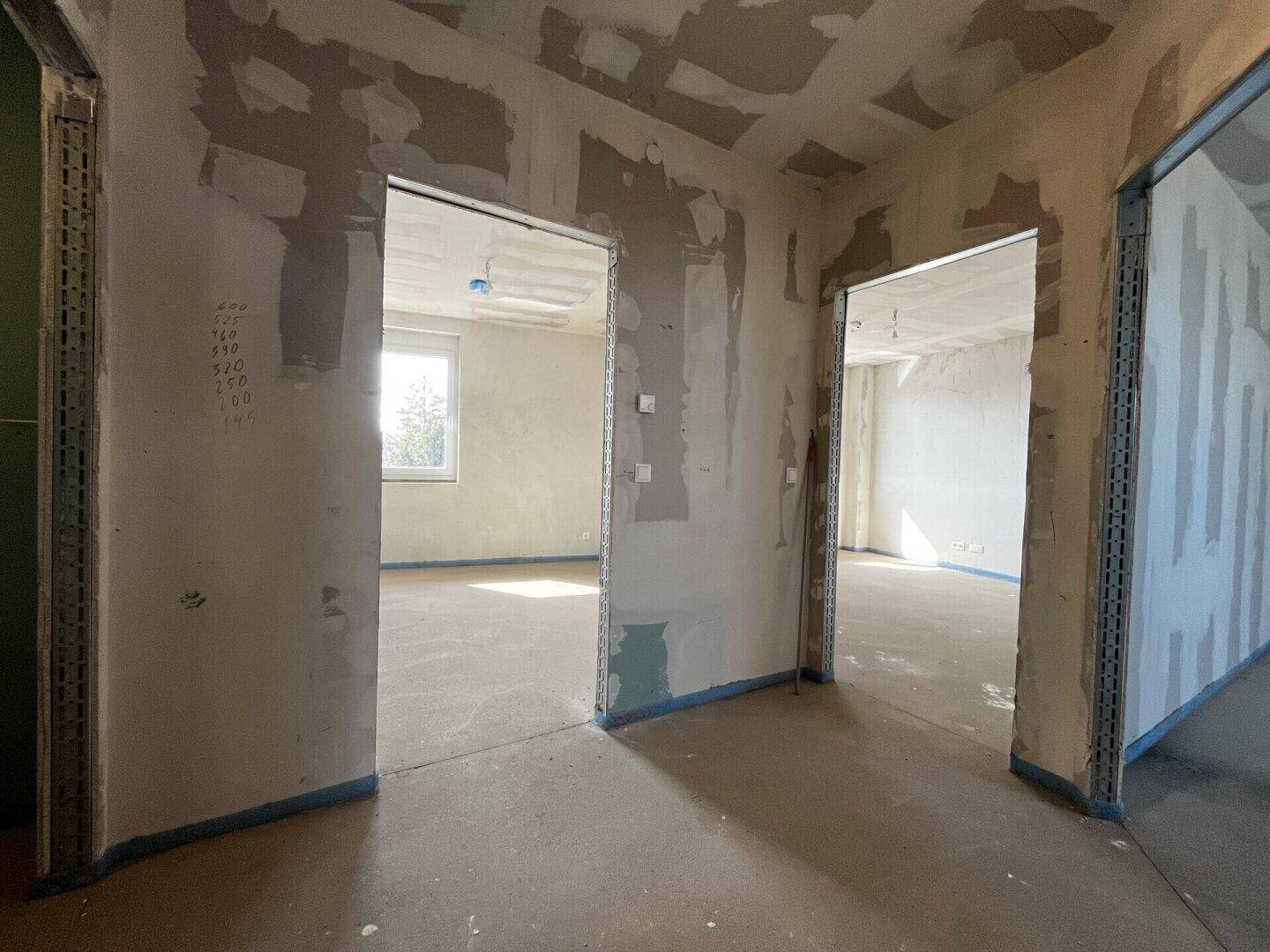 Flur und angrenzende Räume im Rohbau mit Betonboden und Tageslicht.
