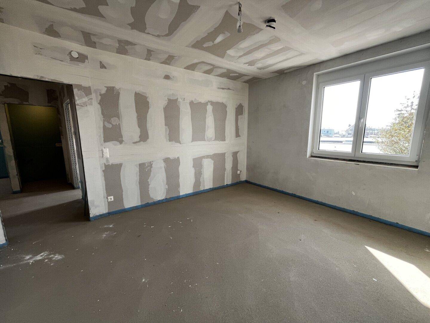 Raum im Rohbau mit Fenster und Blick in einen weiteren unfertigen Bereich.