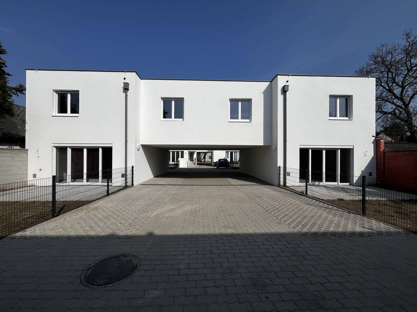 Moderne weiße Gebäudefassade mit großen Fenstern und einem gepflasterten Durchgang, der zu weiteren Gebäuden führt.