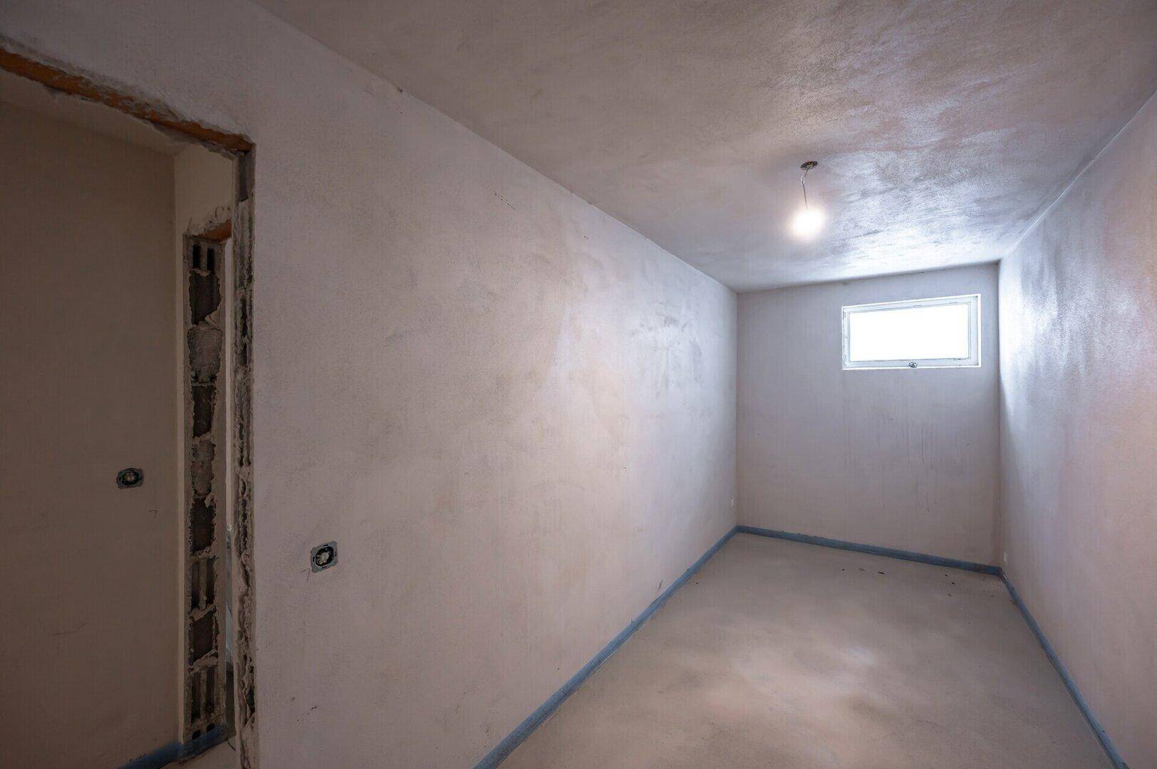 Kleiner Rohbau-Raum mit einem Fenster und Betonboden, geeignet als Abstellraum oder Gästezimmer.