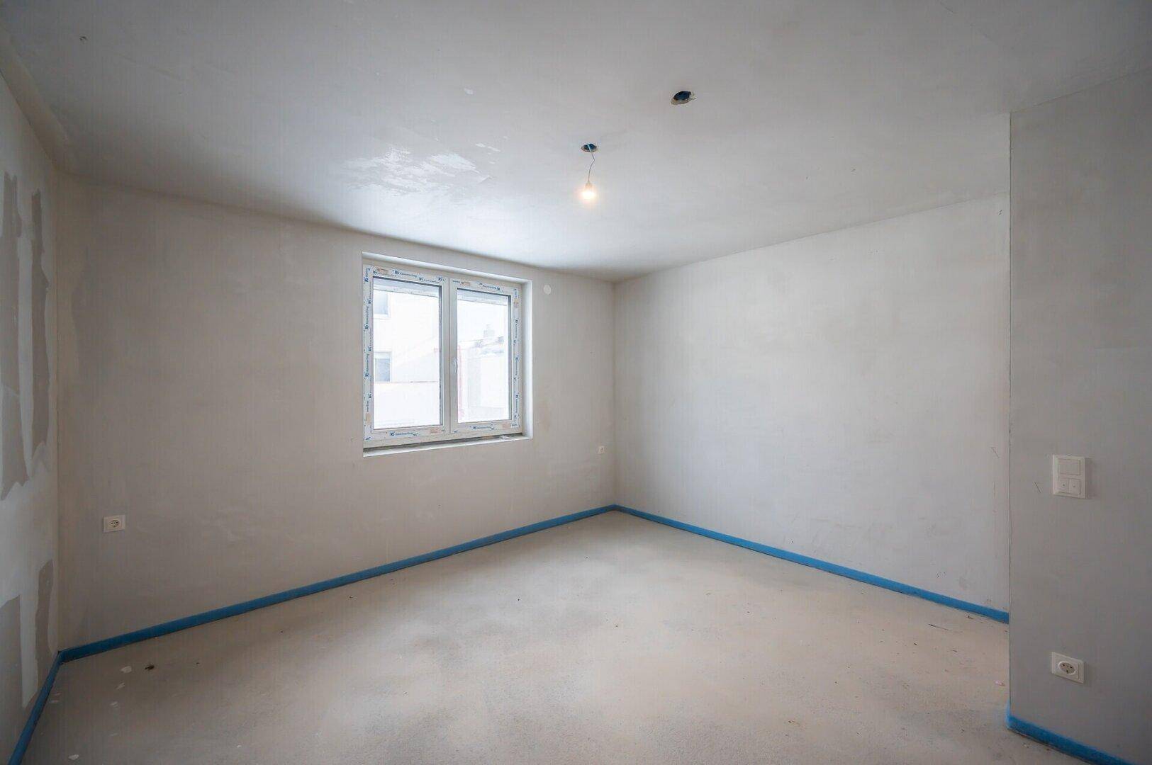 Geräumiger Rohbau-Raum mit Fenster und Betonboden, bereit für den weiteren Innenausbau.