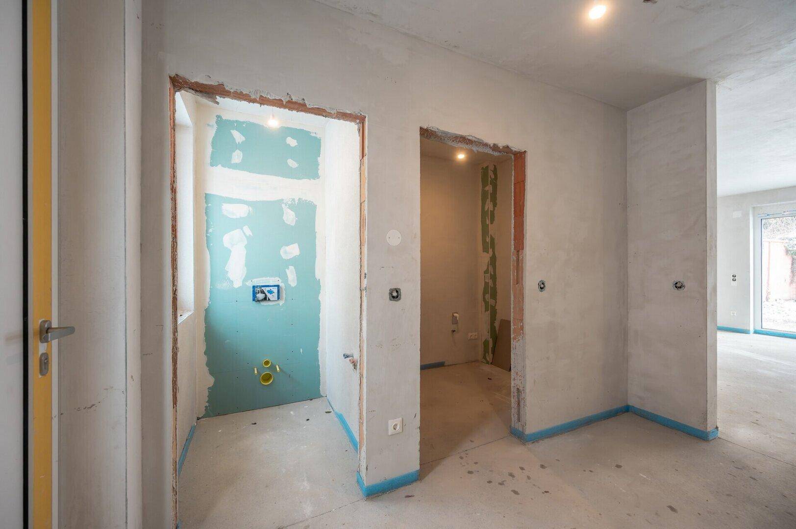 Rohbau-Innenraum mit vorbereiteten Türöffnungen und sichtbaren Sanitärinstallationen für ein Badezimmer.