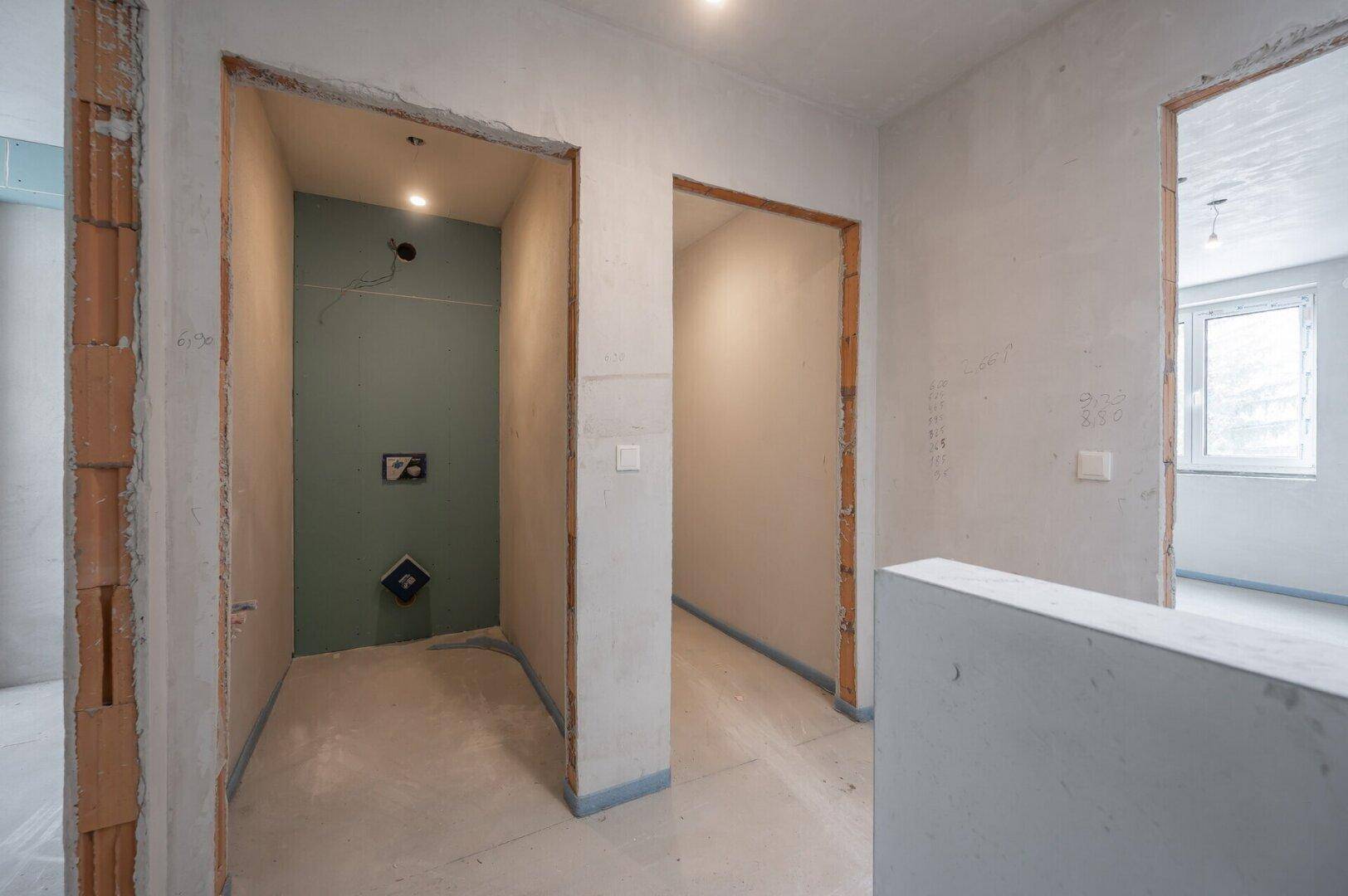 Rohbau-Flur mit vorbereiteten Türöffnungen und sichtbaren Installationen für ein zukünftiges Badezimmer.
