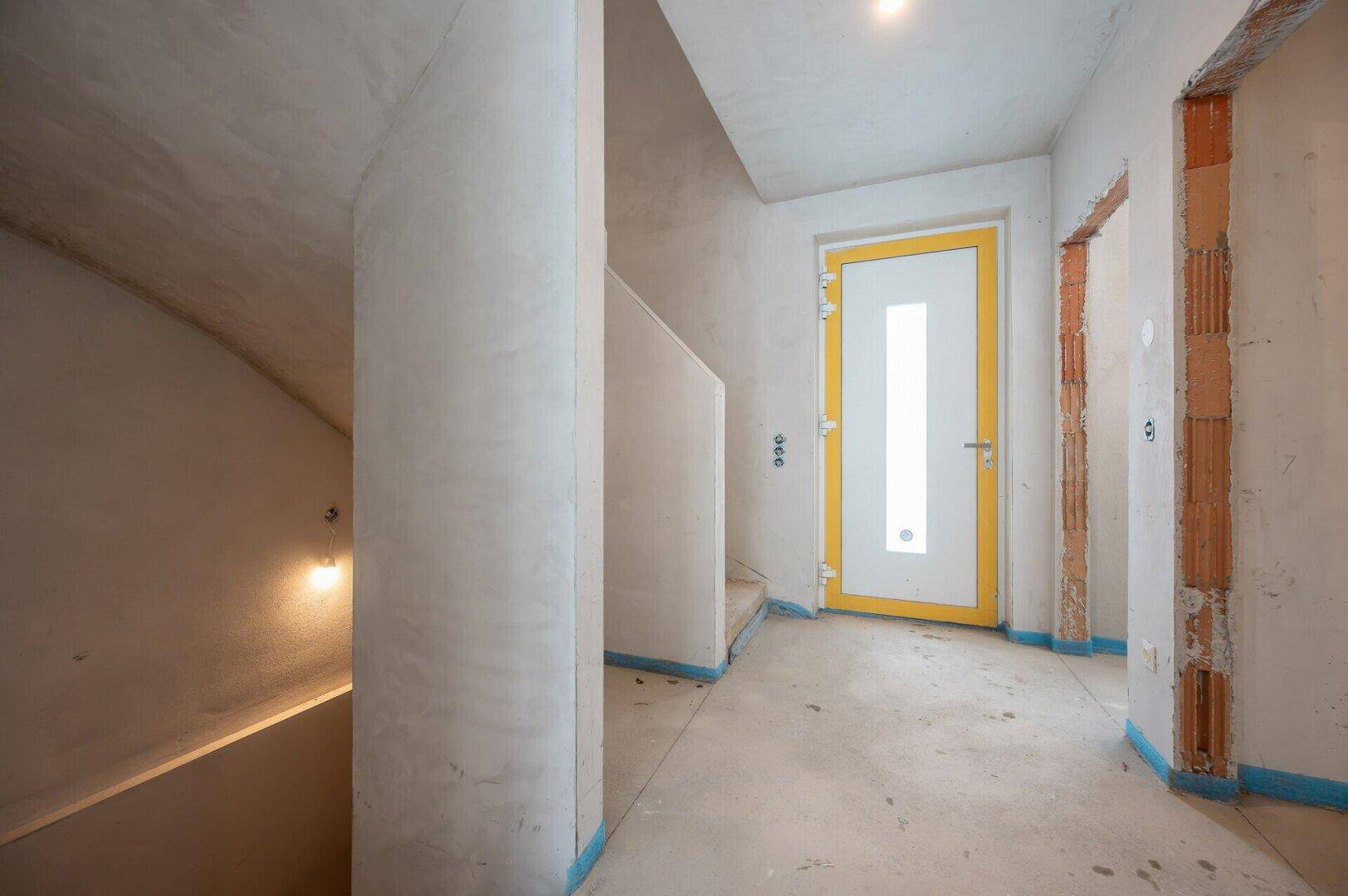 Unfertiger Eingangsbereich mit Rohbau-Treppe und einer Tür mit gelbem Rahmen.