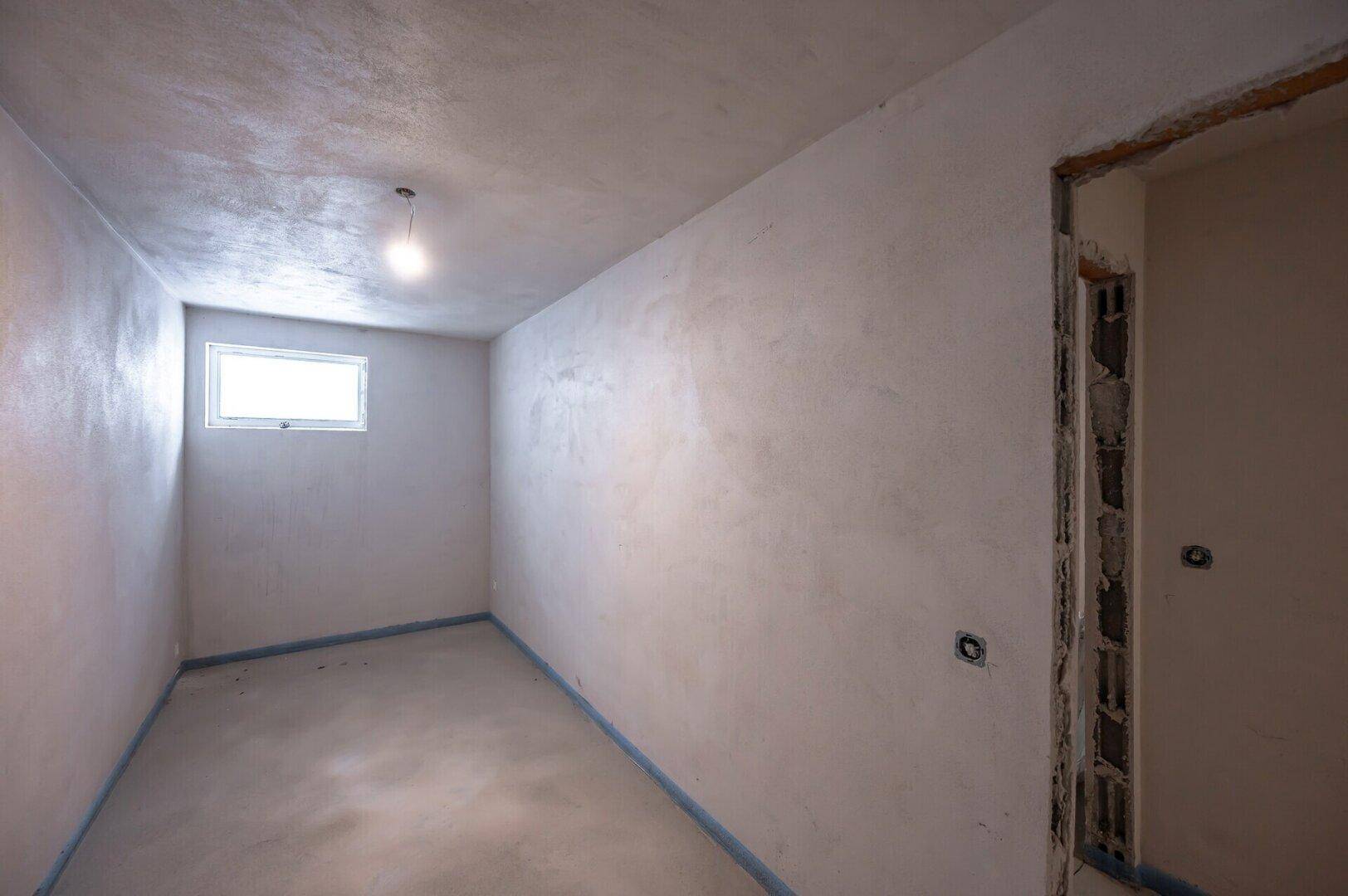Kleiner, unfertiger Raum im Rohbau mit einem hochgelegenen Fenster, ideal als Abstellraum.