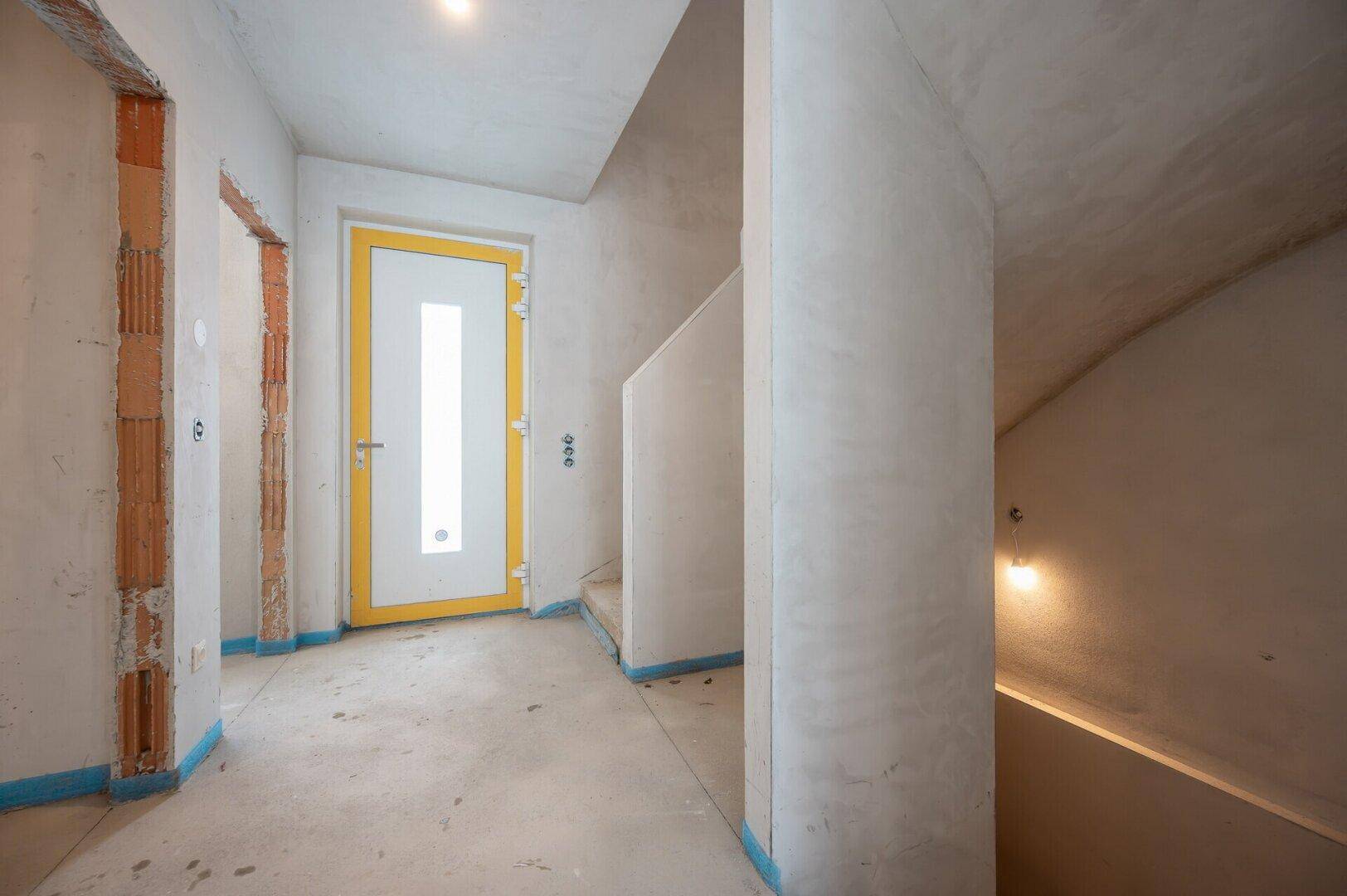 Unfertiger Flur im Rohbau mit gelb gerahmter Eingangstür und rohen Betontreppen.