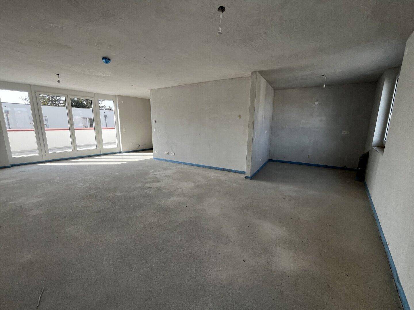 Geräumiger Rohbau-Wohnbereich mit Betonboden und großen Fensterfronten, die viel Licht hereinlassen.