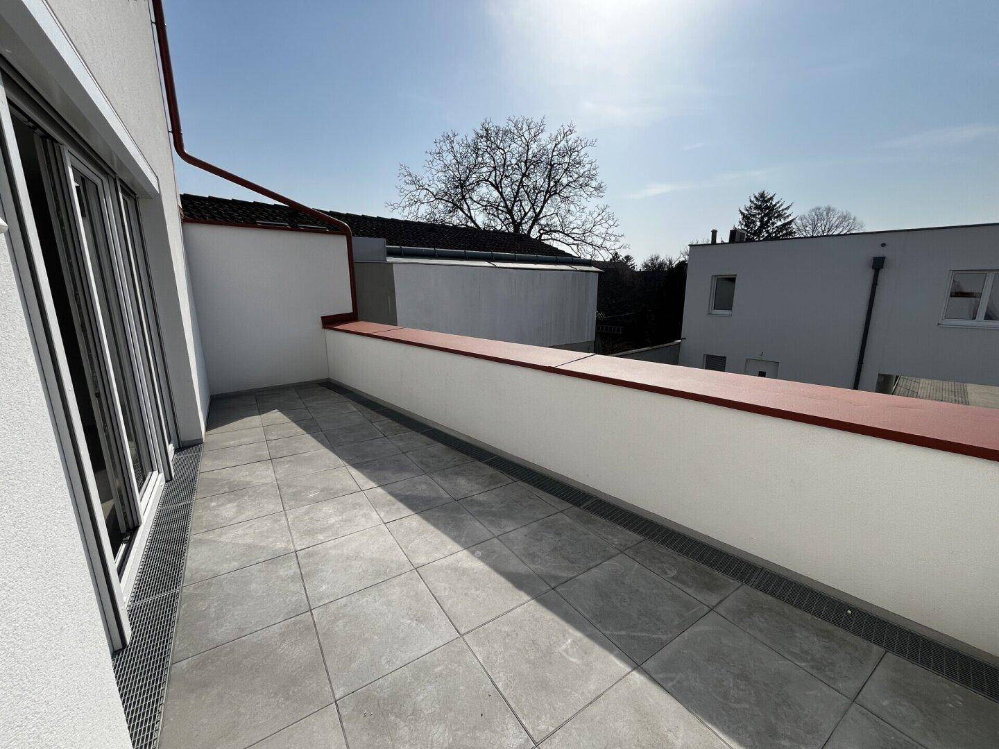Leere, sonnige Terrasse mit Fliesenboden und weißer Brüstung, bietet Platz für individuelle Gestaltung.