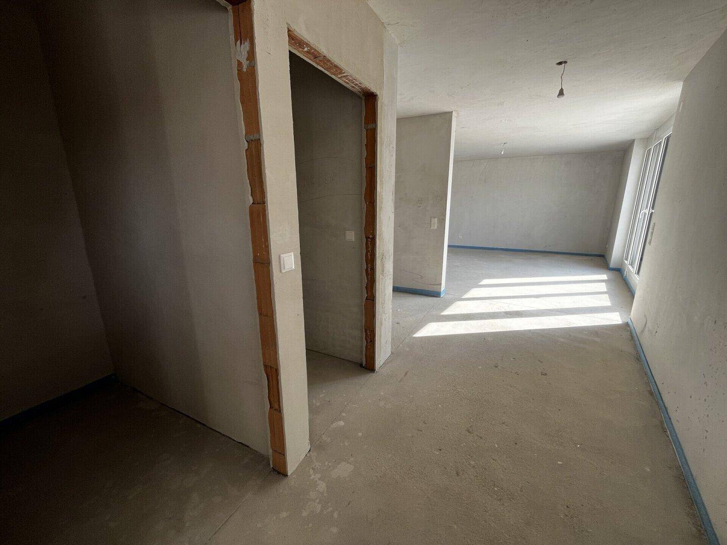 Unfertiger Innenraum im Rohbau mit Betonboden und viel natürlichem Licht durch die Fenster.