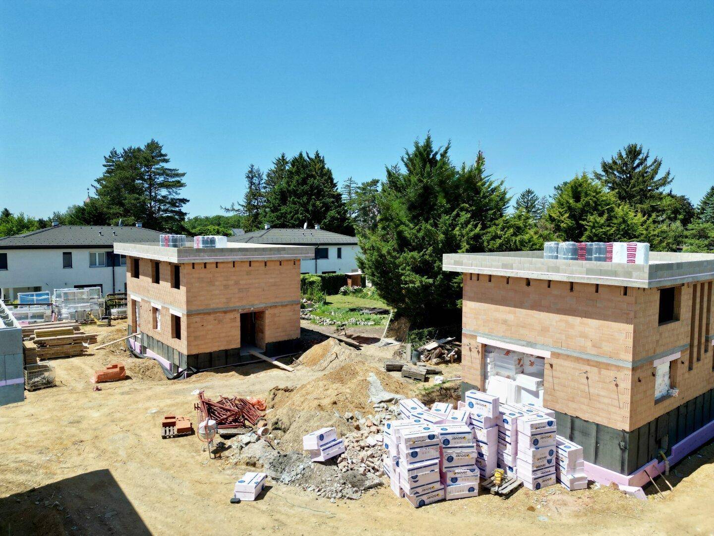 Zwei Neubauhäuser im Rohbau auf einer sonnigen Baustelle mit Baumkulisse im Hintergrund.