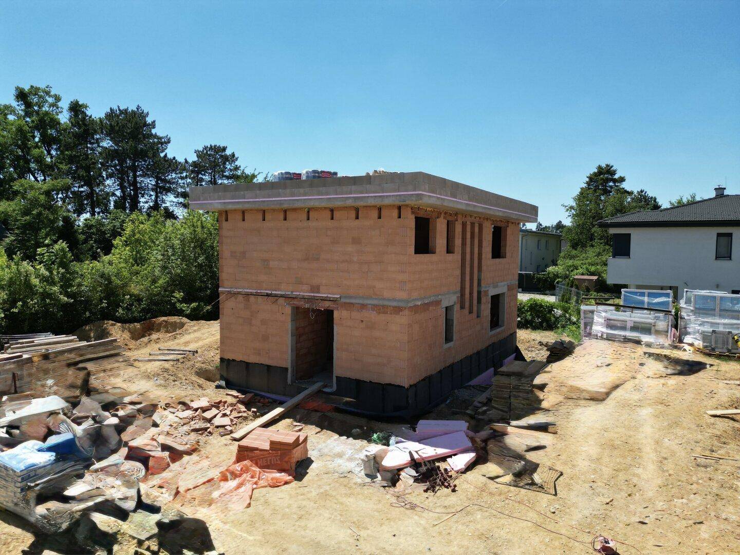 Einzelnes Haus im Rohbau auf einer Baustelle, umgeben von Bäumen und Baumaterialien.