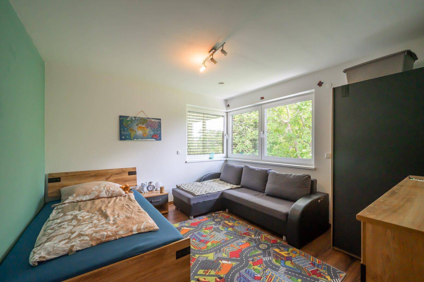 Helles Schlafzimmer mit Einzelbett, Sofa und Blick ins Grüne durch große Fenster.