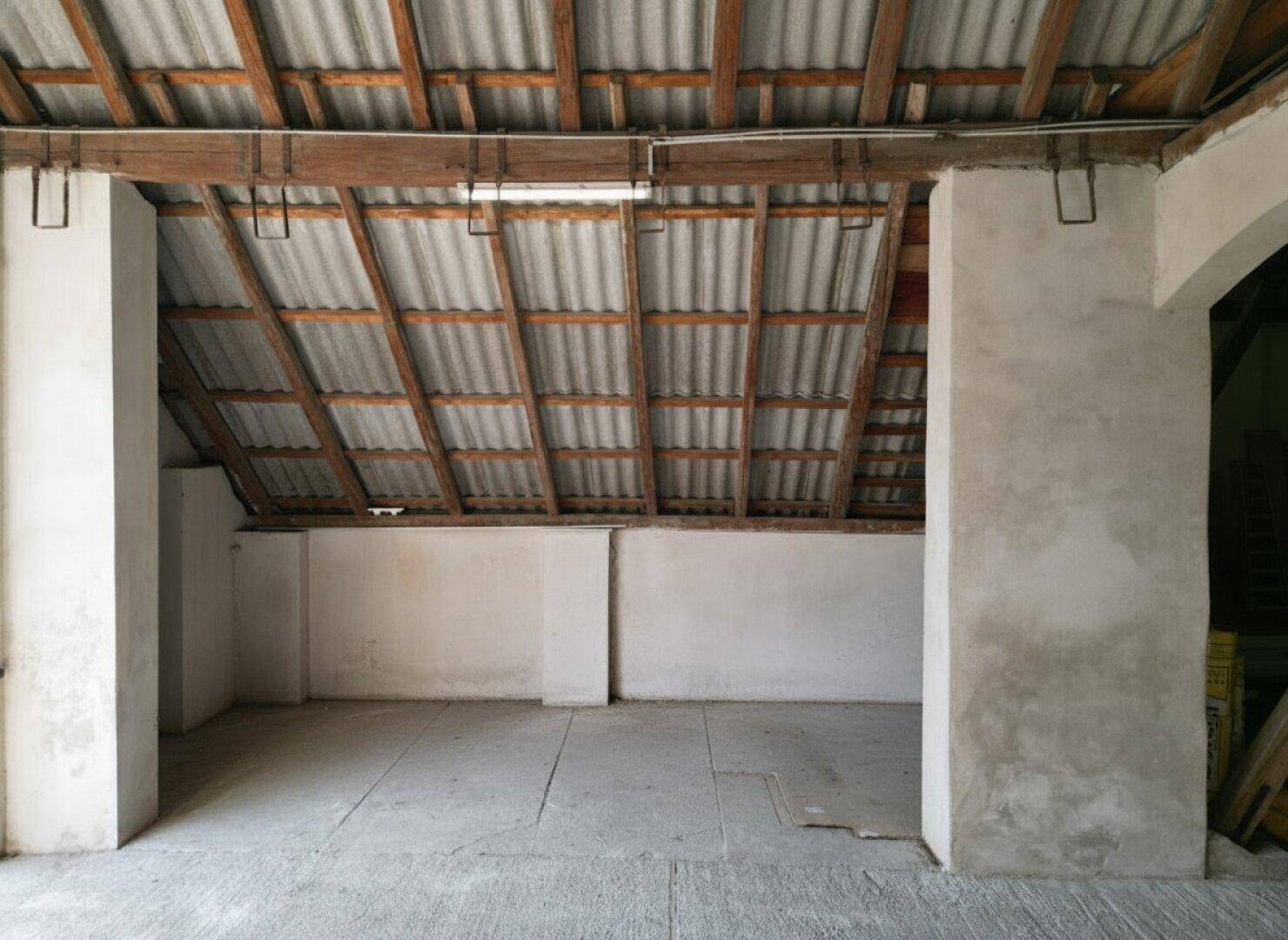 Innenraum mit schräger Decke, freiliegenden Dachsparren und einem einfachen Betonboden.