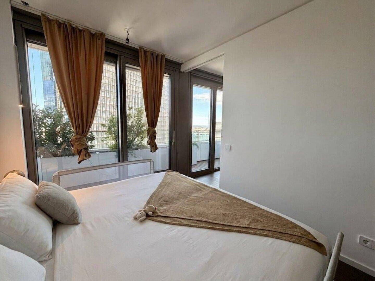 Helles Schlafzimmer mit großen Fenstern, Vorhängen und Blick auf die Stadt.