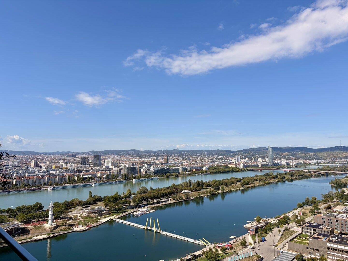 Panoramablick auf die Stadt Wien mit der Donau und umliegenden Hügeln unter blauem Himmel.