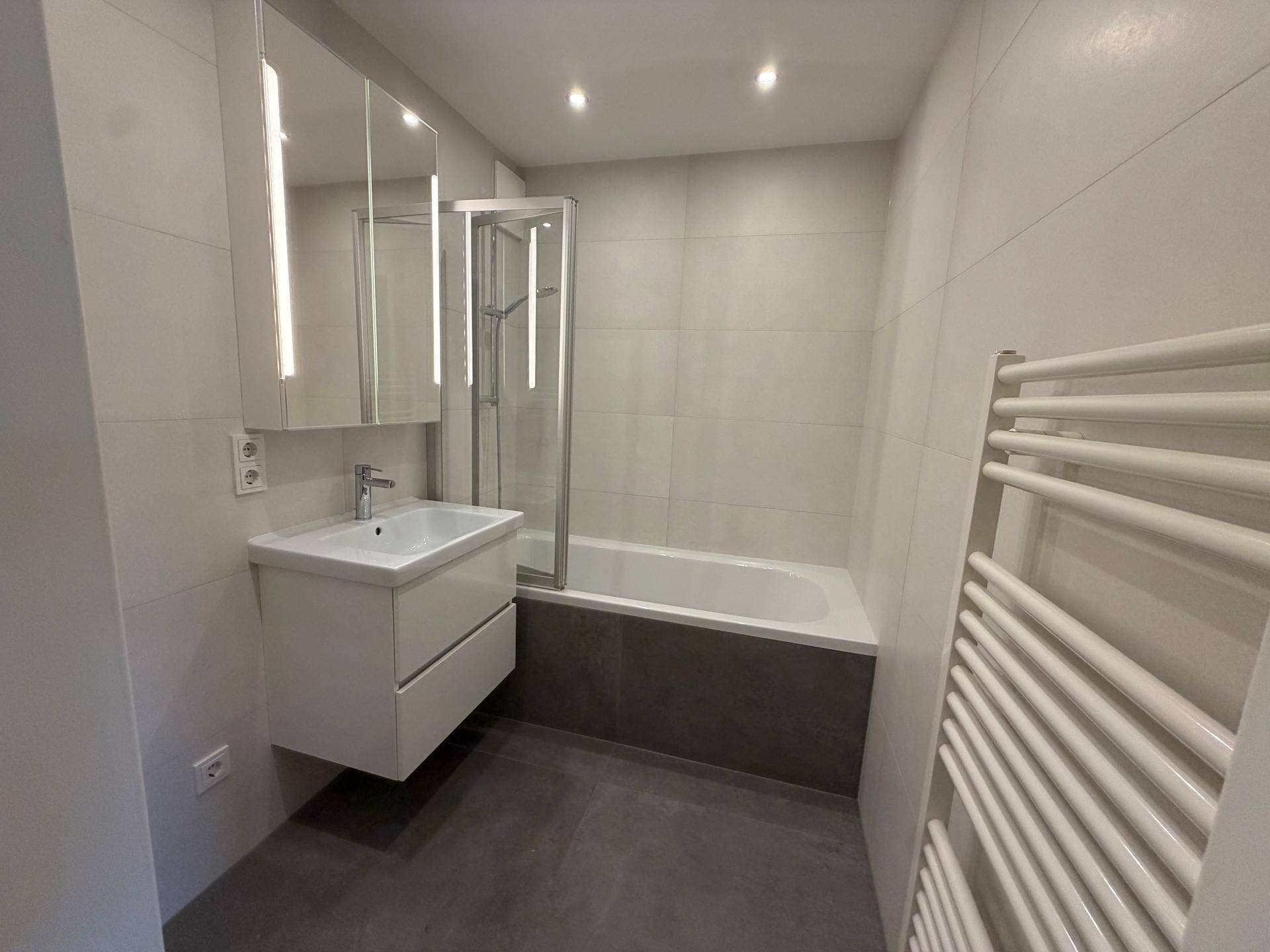 Modernes Badezimmer mit weißen Fliesen, schwebendem Waschtisch, Badewanne mit Duschwand und Handtuchheizkörper.