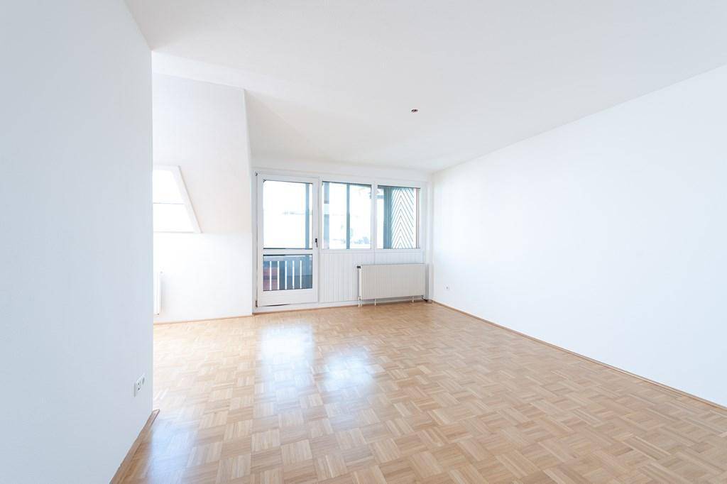 Heller, leerer Wohnraum mit Parkettboden, weißen Wänden und Zugang zu einem kleinen Balkon.