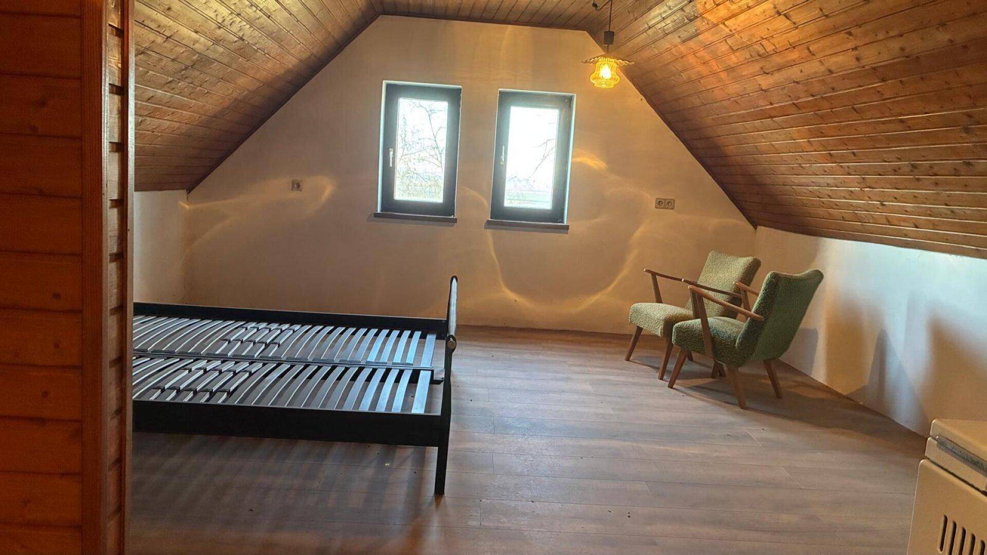 Geräumiges Dachgeschoss-Schlafzimmer mit Holzbalkendecke, zwei Fenstern und Sitzgelegenheiten.