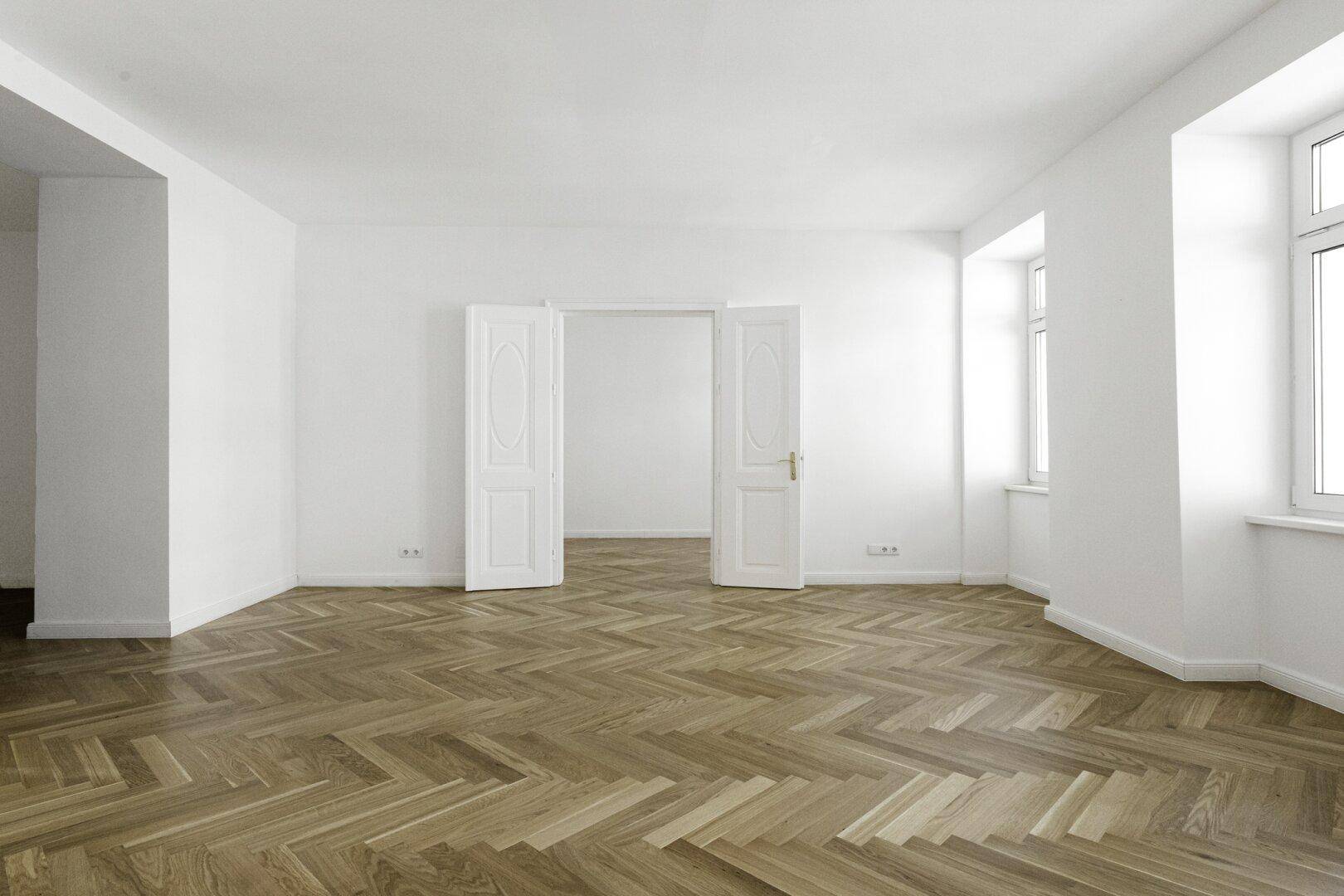 Geräumiger Wohnbereich mit klassischem Fischgrätparkett und doppelten Flügeltüren zu einem weiteren Zimmer.